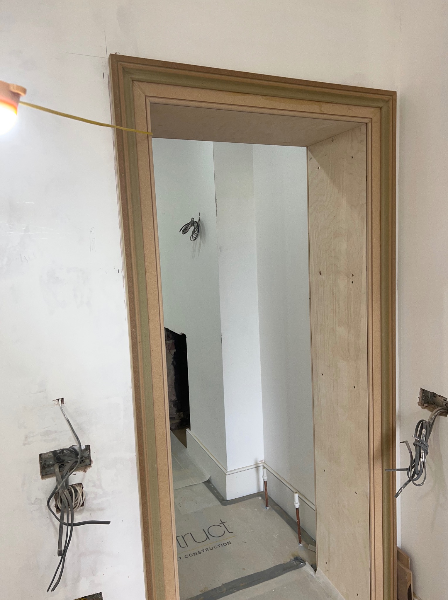 Surowa, drewniana rama drzwiowa w trakcie montażu, widoczne przewody elektryczne w ścianie. Wnętrze w stanie deweloperskim, przygotowane do wykończenia.