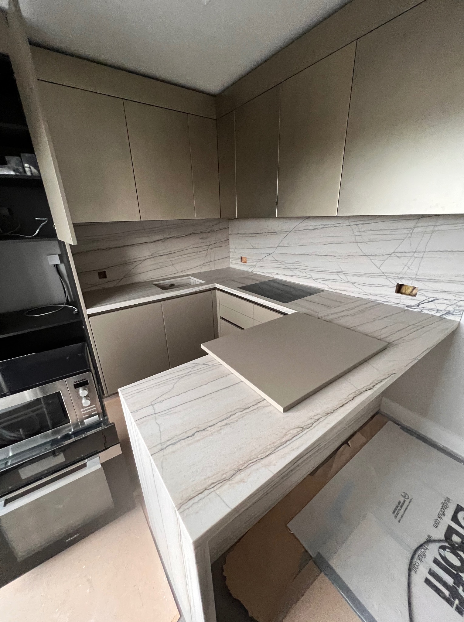 Nowoczesna kuchnia z beżowymi szafkami i blatem z marmurowym wzorem. Widoczny fragment płyty indukcyjnej i zlewu. Na blacie leży płyta.