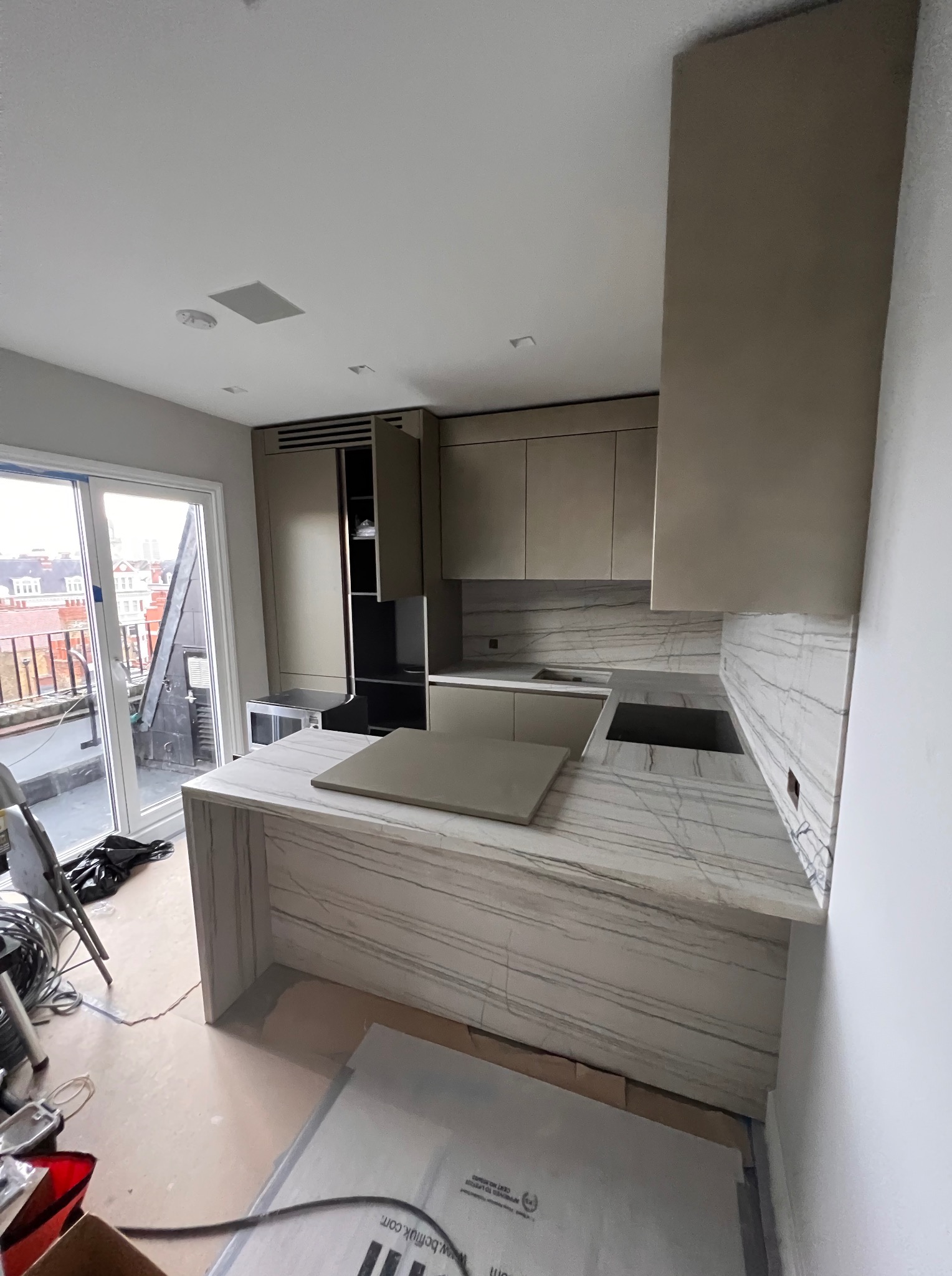 Jasna kuchnia z wyspą w trakcie montażu, widok na balkon. Marmurowy blat i okładzina ściany, minimalistyczne szafki. Elementy instalacji widoczne na podłodze.