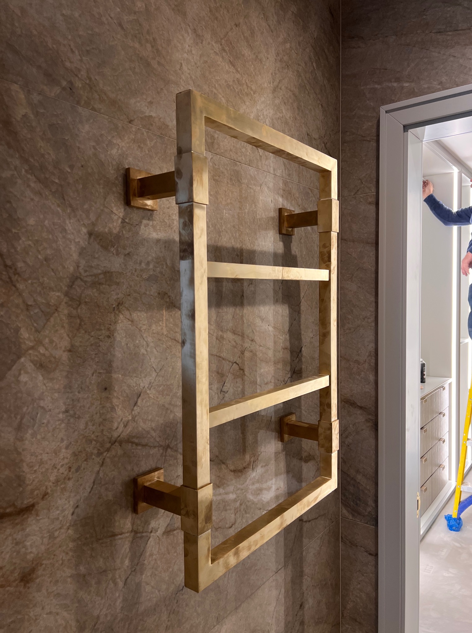 Złoty, kwadratowy grzejnik łazienkowy na marmurowej ścianie. Widoczny fragment wnętrza łazienki z szafkami i drabina. Ujęcie z bliska, prezentujące detale wykończenia.