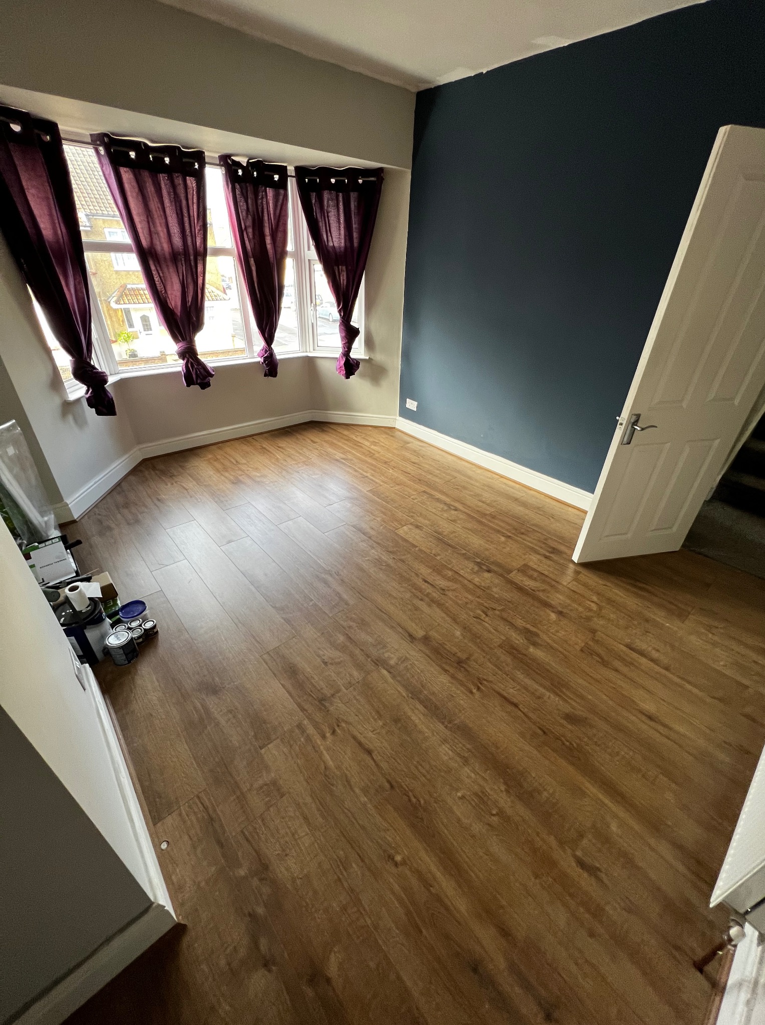 Pokój po remoncie: drewniana podłoga, granatowa ściana, okno z fioletowymi zasłonami. Widoczne puszki z farbą, sugerujące świeżo zakończone prace malarskie.