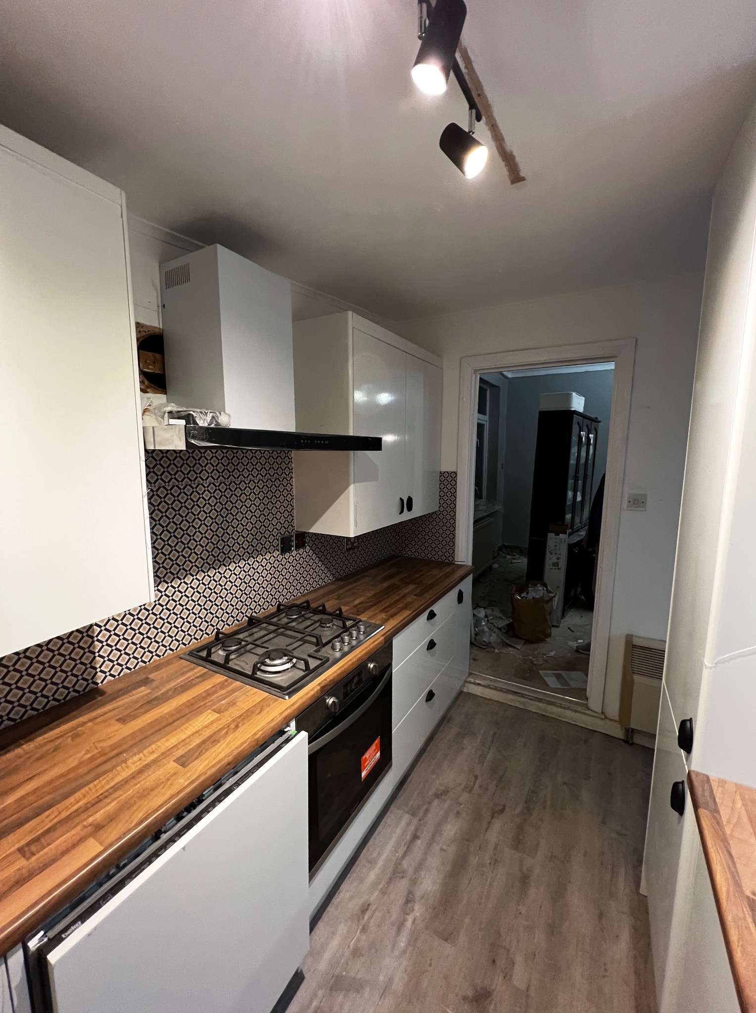 Biała kuchnia z drewnianym blatem i wzorzystą mozaiką nad kuchenką. Widoczny fragment pomieszczenia w trakcie remontu.