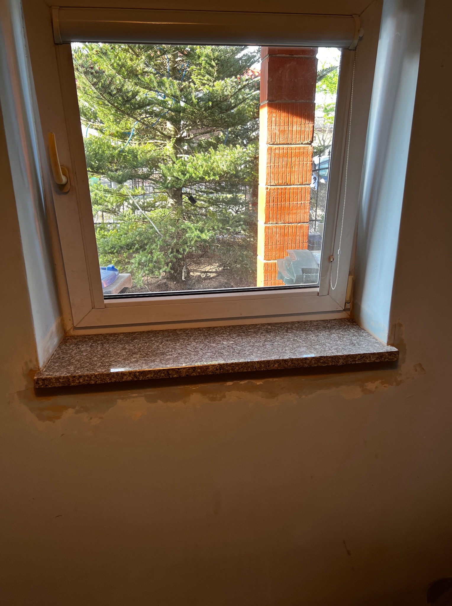 Granitowy parapet wewnętrzny pod oknem dachowym, widok na zieleń drzew. Ślady po pracach wykończeniowych na ścianie. Jasne, naturalne światło.