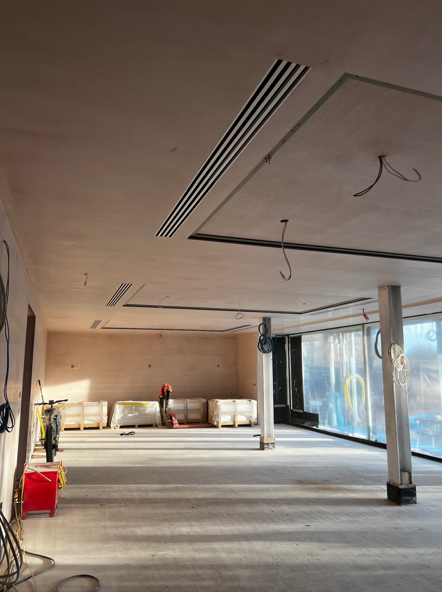 Wnętrze w trakcie wykończenia: gładzie na ścianach, kratki wentylacyjne w suficie, folia ochronna na oknach. Widoczny pracownik w oddali i materiały budowlane.