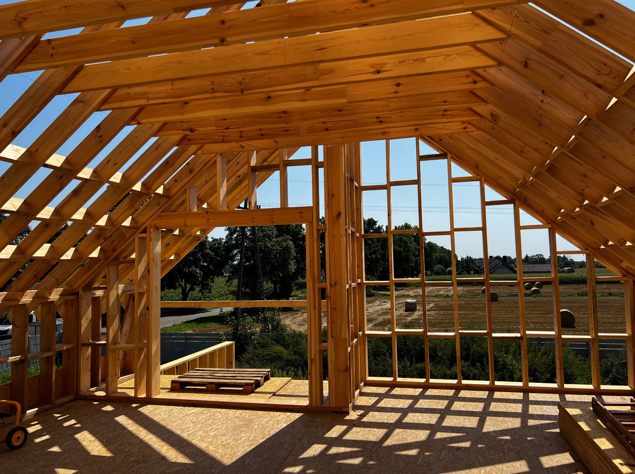 Drewniana konstrukcja dachu w trakcie budowy, widok z wnętrza. Elementy więźby dachowej z jasnego drewna na tle błękitnego nieba i krajobrazu z polami.