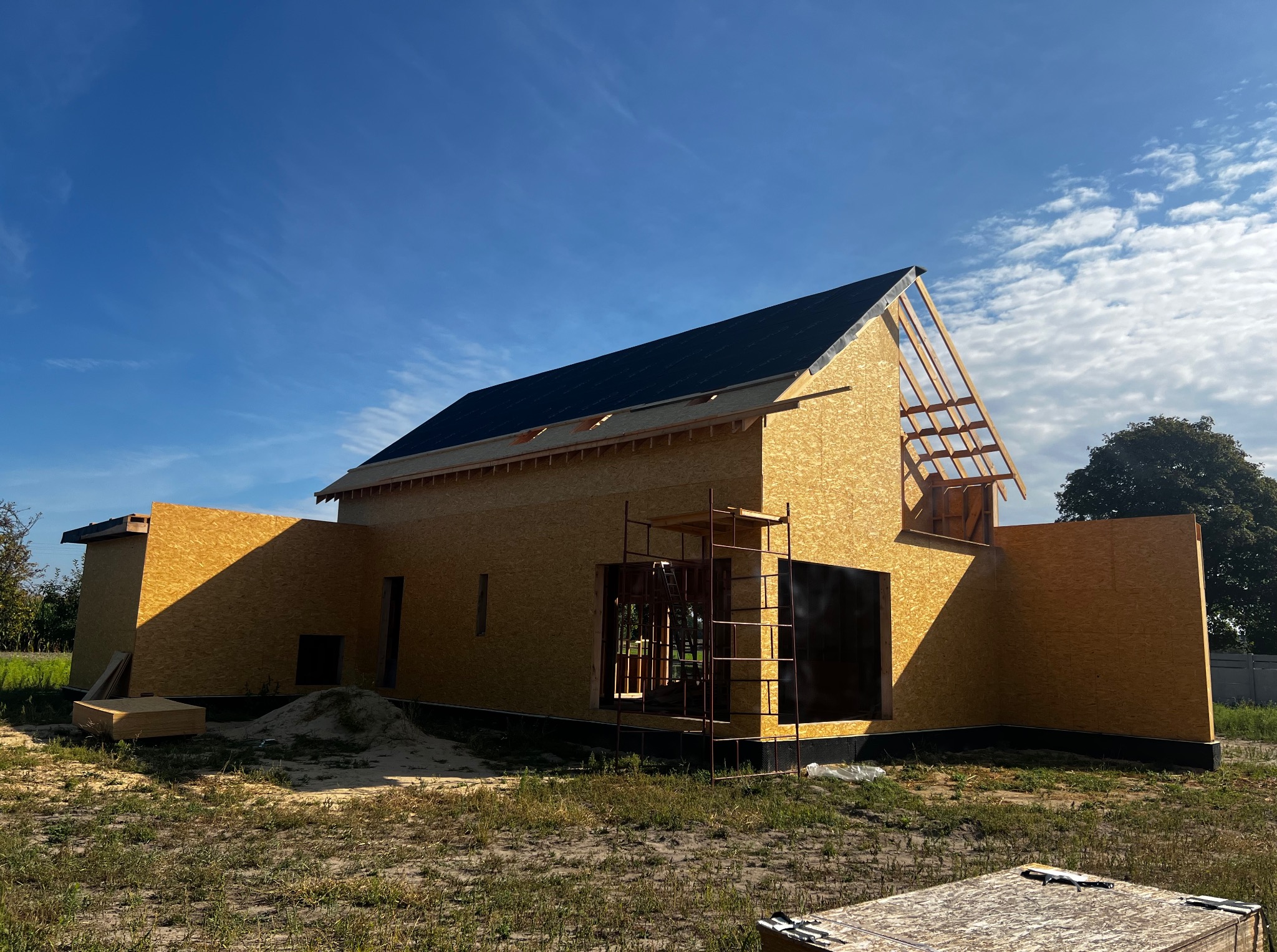 Dom w trakcie budowy: konstrukcja z płyt OSB, częściowo pokryty dach, rusztowanie przy oknie. Widok na budynek w słoneczny dzień.