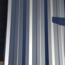 P.H.U. JUDYTA KUCZYŃSKA - Błyszcząca, srebrna blacha trapezowa z wyraźnym cieniem, ułożona pionowo. Widoczne regularne przetłoczenia i odbicia światła. Fragment podłoża w lewym dolnym rogu.