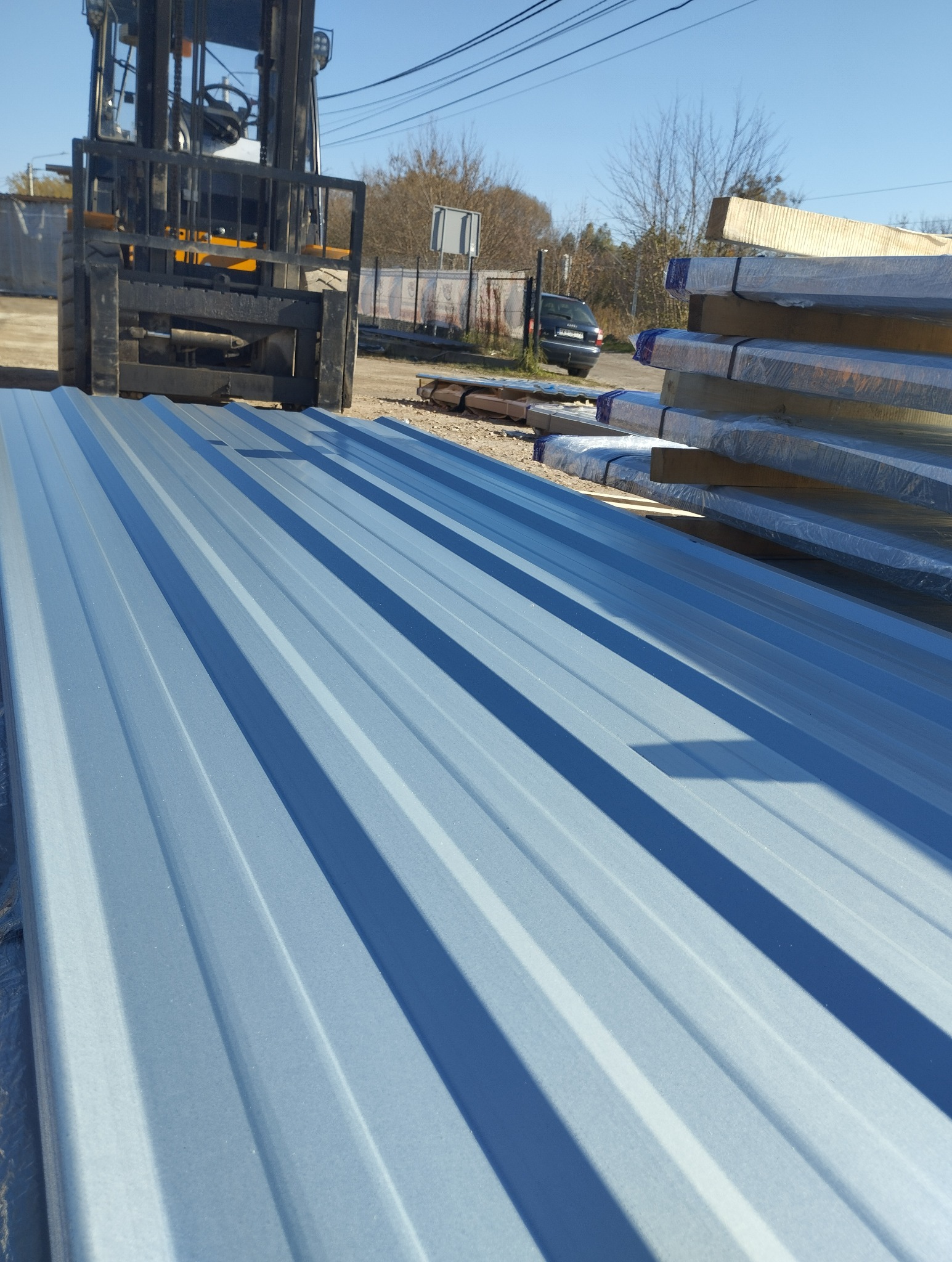 Jasnoszara blacha trapezowa na placu budowy, widoczny wózek widłowy i zapakowane elementy. Słoneczny dzień, błękitne niebo w tle.