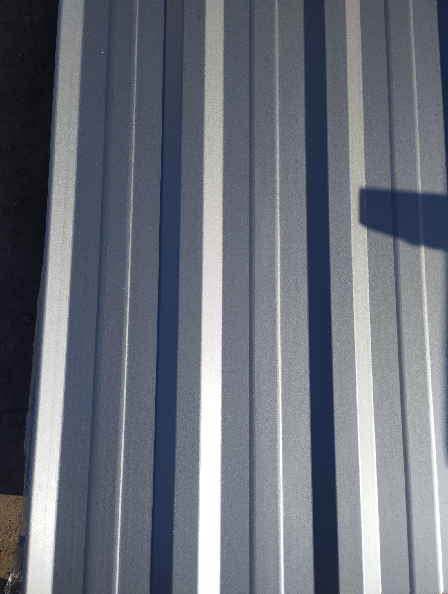 Błyszcząca, srebrna blacha trapezowa z wyraźnym cieniem, ułożona pionowo. Widoczne regularne przetłoczenia i odbicia światła. Fragment podłoża w lewym dolnym rogu.