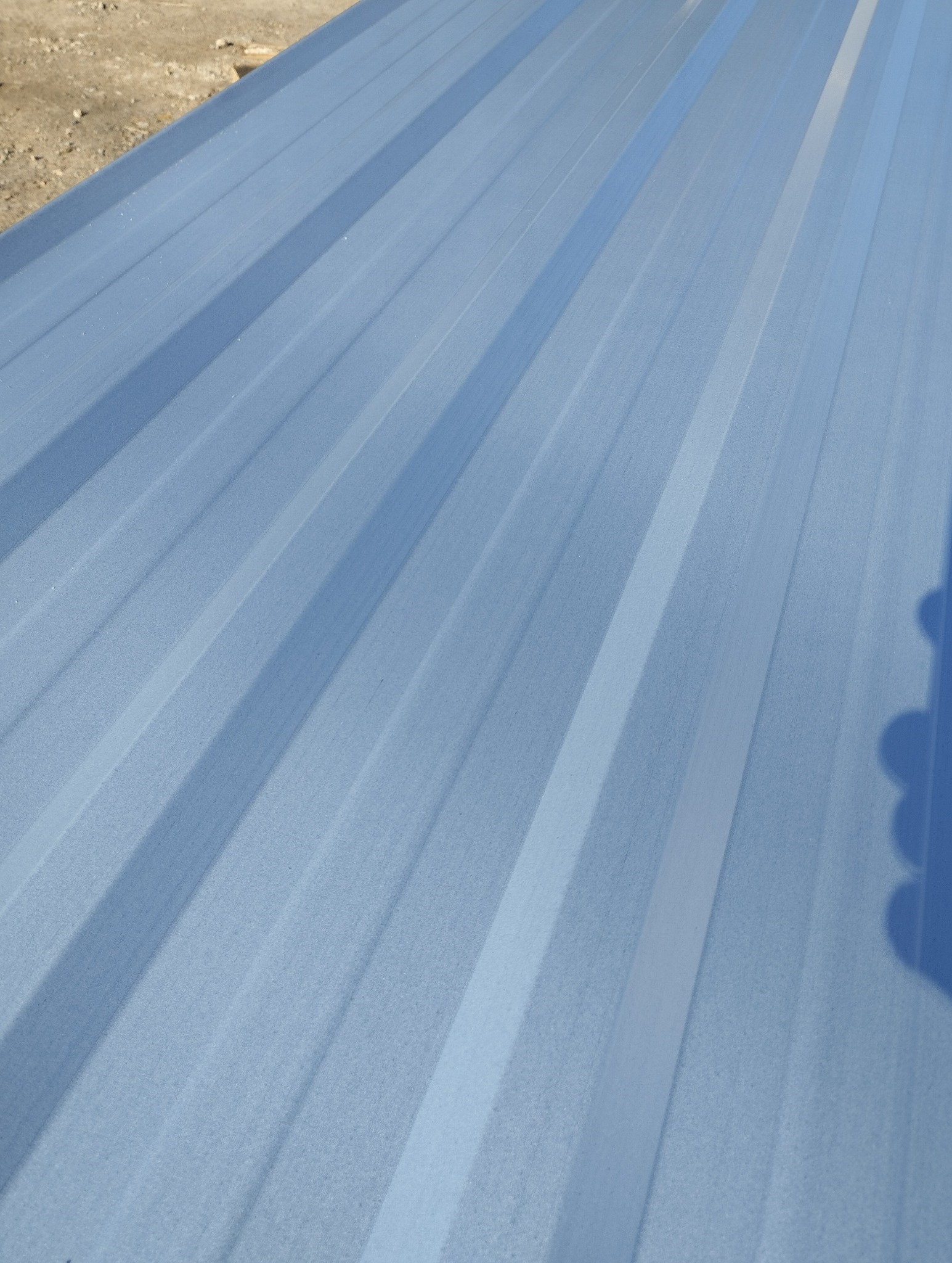 Jasnoniebieska blacha trapezowa z widocznym cieniem, ułożona pod kątem, z fragmentem ziemi w tle. Widoczne delikatne refleksy światła na powierzchni.
