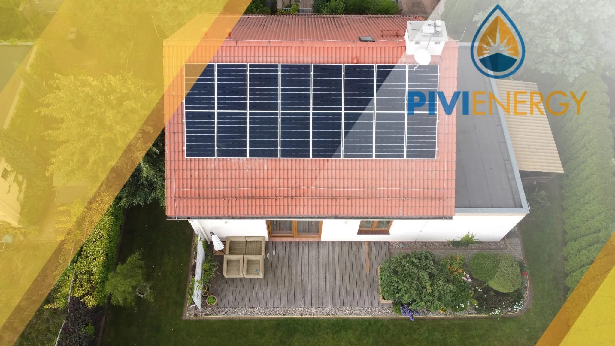 Widok z góry na dom z czerwoną dachówką i panelami słonecznymi. Na dachu widoczna instalacja fotowoltaiczna. Obok domu ogród i taras z meblami.