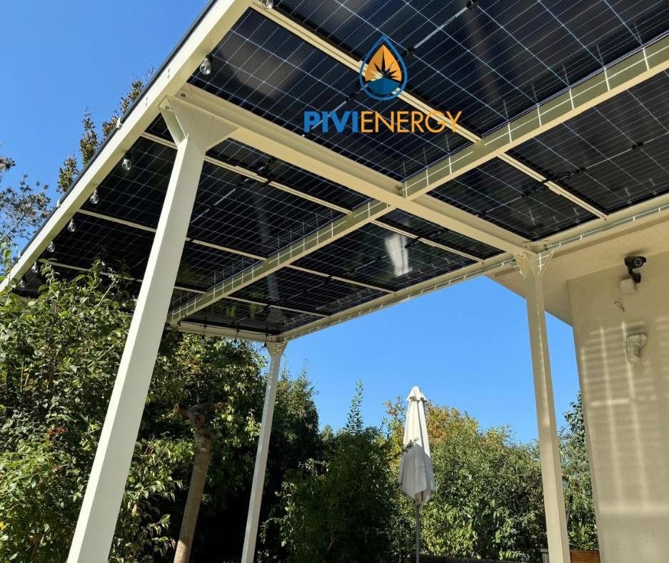 Zadaszenie tarasu z panelami słonecznymi w słoneczny dzień. Widoczne białe podpory konstrukcji i logo firmy na panelach. Tło: zieleń drzew i błękitne niebo.