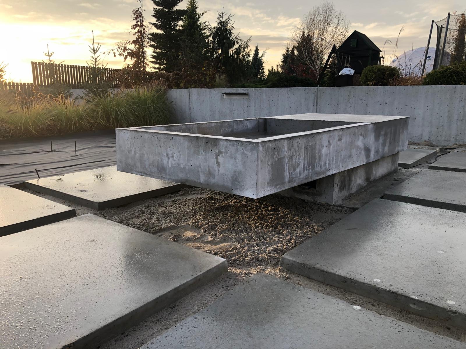 Betonowa konstrukcja w ogrodzie w trakcie budowy, z widocznymi płytami chodnikowymi i przygotowanym podłożem, w tle zieleń i zachodzące słońce.