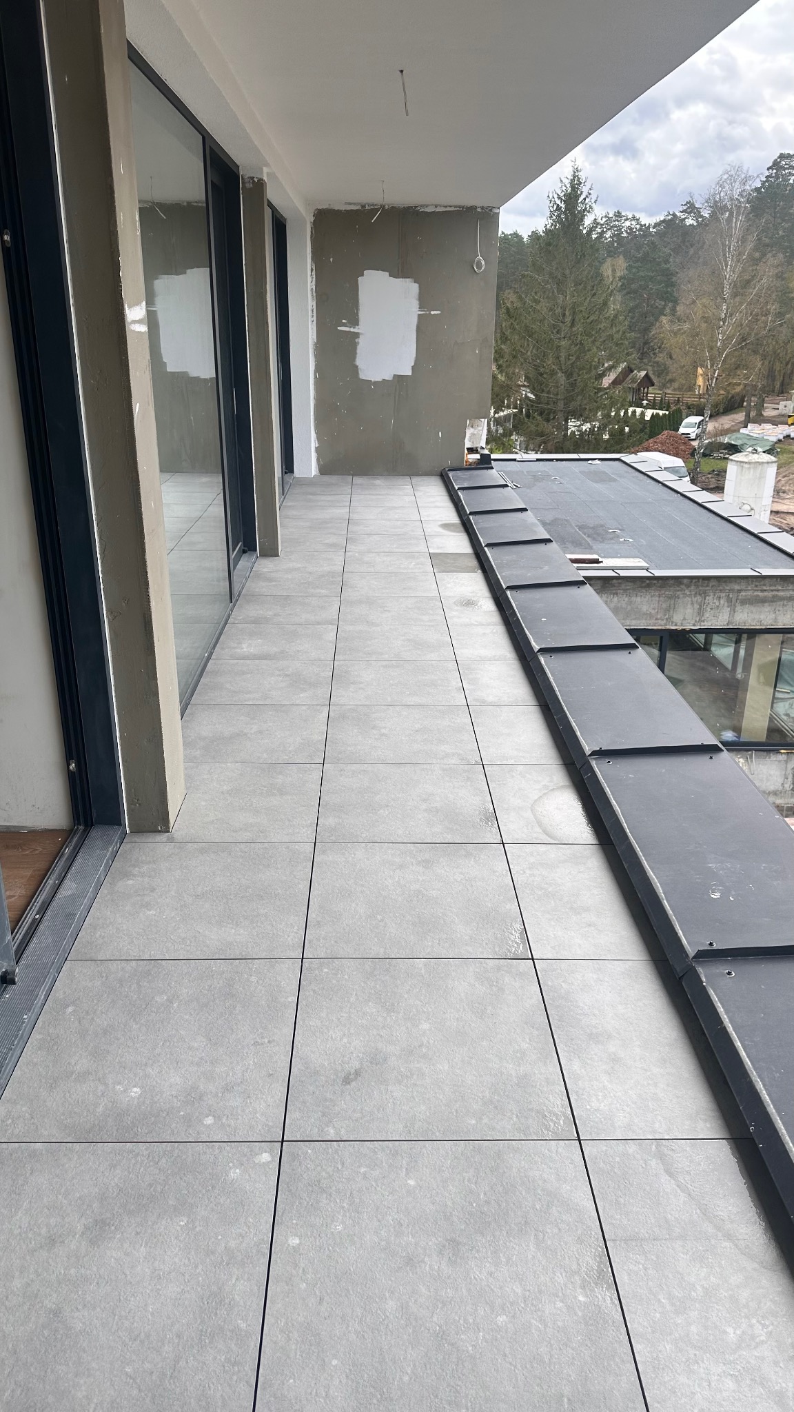Wyłożony szarymi płytkami taras z widokiem na zieleń. Widoczne fragmenty okien i ściany w trakcie prac wykończeniowych. Minimalistyczny styl.