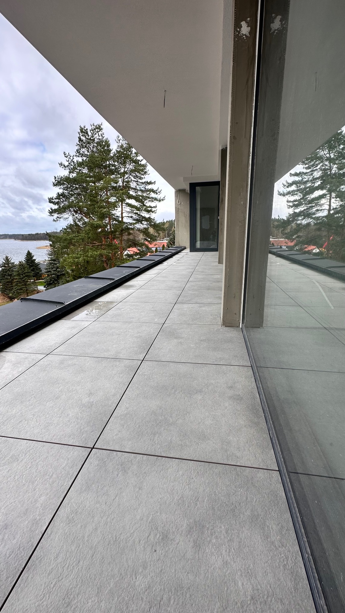 Wyłożony szarymi płytkami taras z widokiem na jezioro i las. Widoczne betonowe filary i duże okno. Minimalistyczny design z elementami natury w tle.