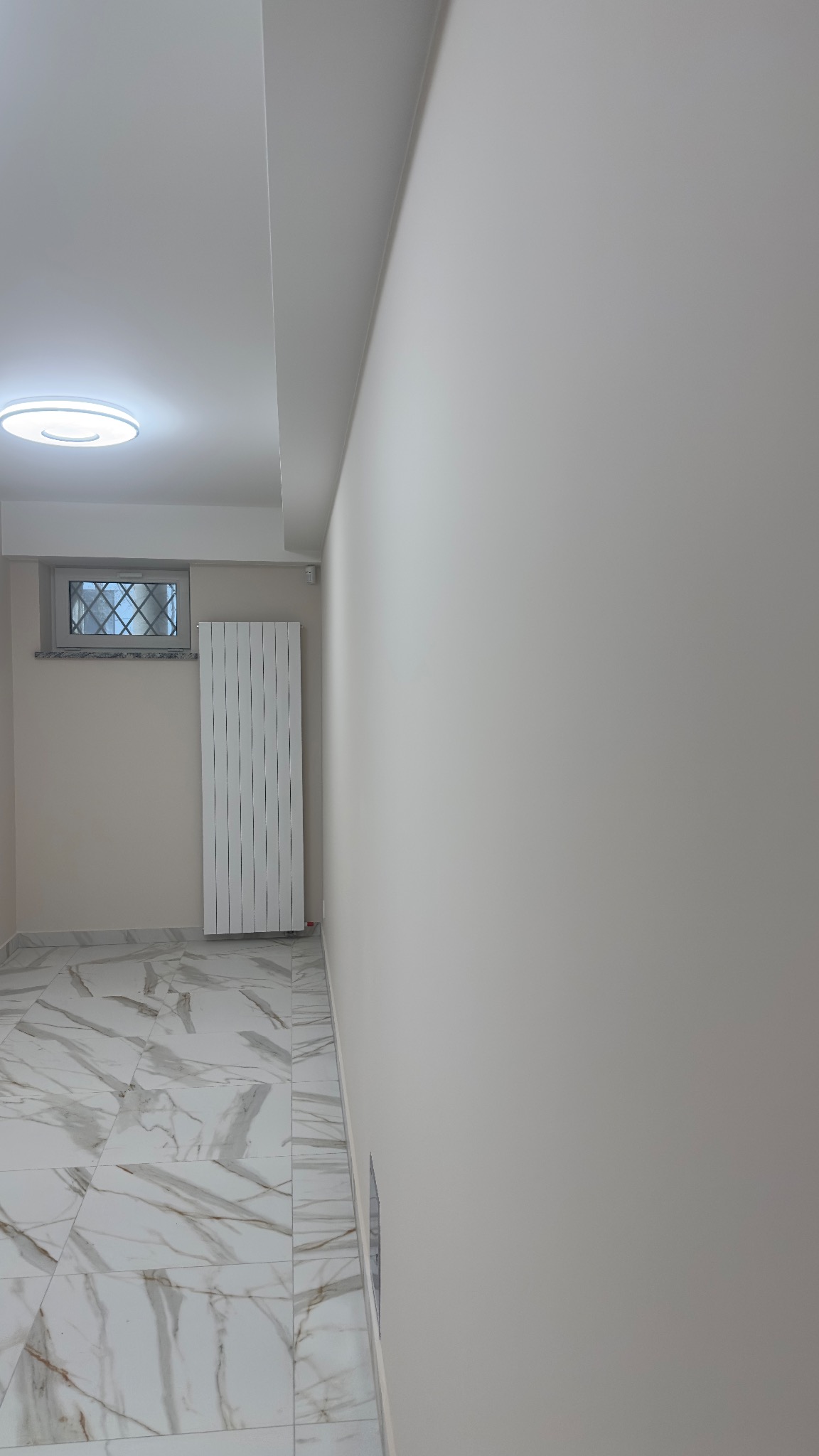 Jasne wnętrze po remoncie: marmurowe płytki podłogowe, świeżo pomalowane ściany w odcieniach bieli, grzejnik panelowy i okno z ozdobną kratą. Minimalistyczny design.