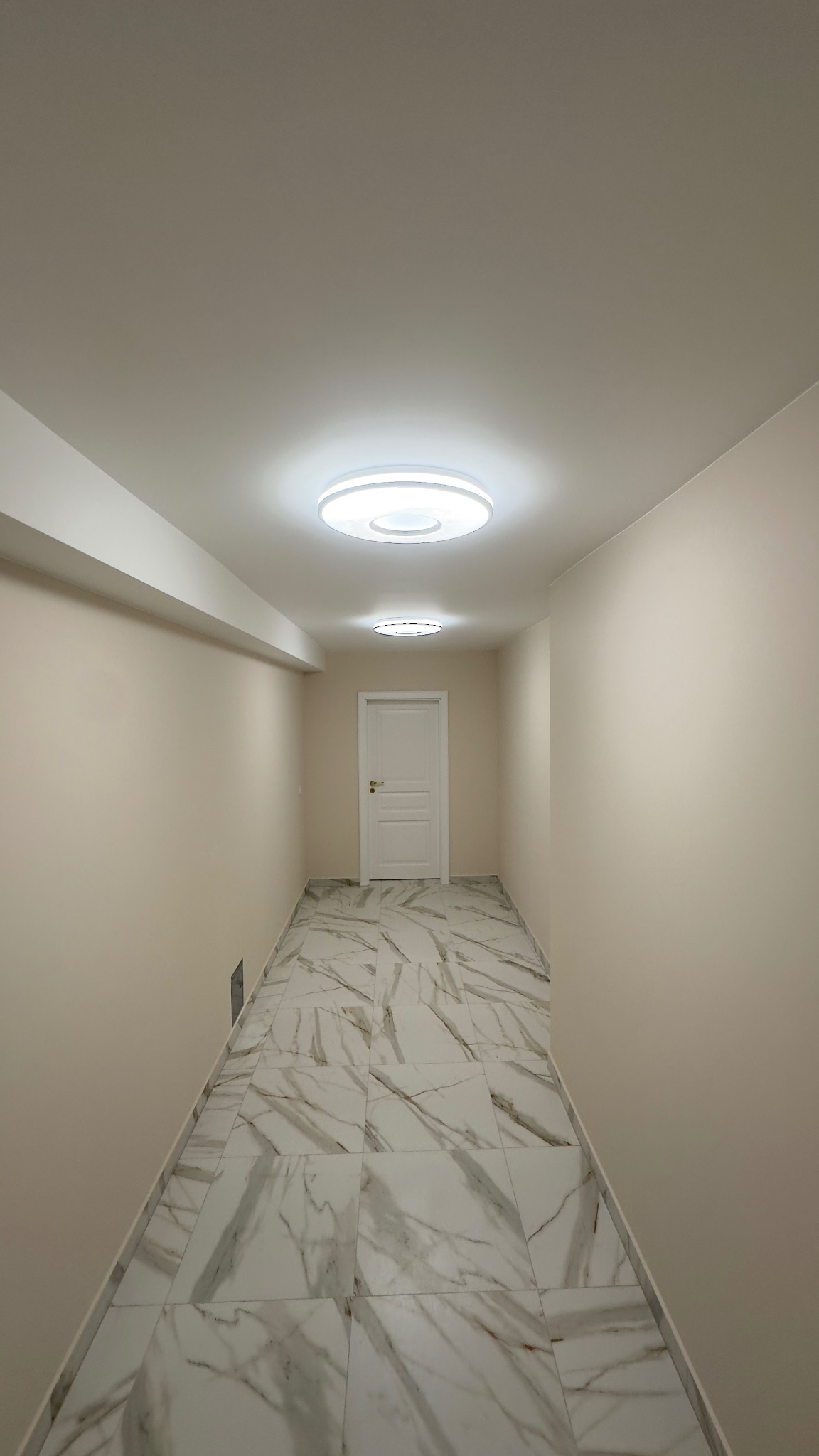 Korytarz po remoncie z białymi ścianami, marmurową podłogą i nowoczesnym oświetleniem sufitowym. Widok na białe drzwi na końcu korytarza.