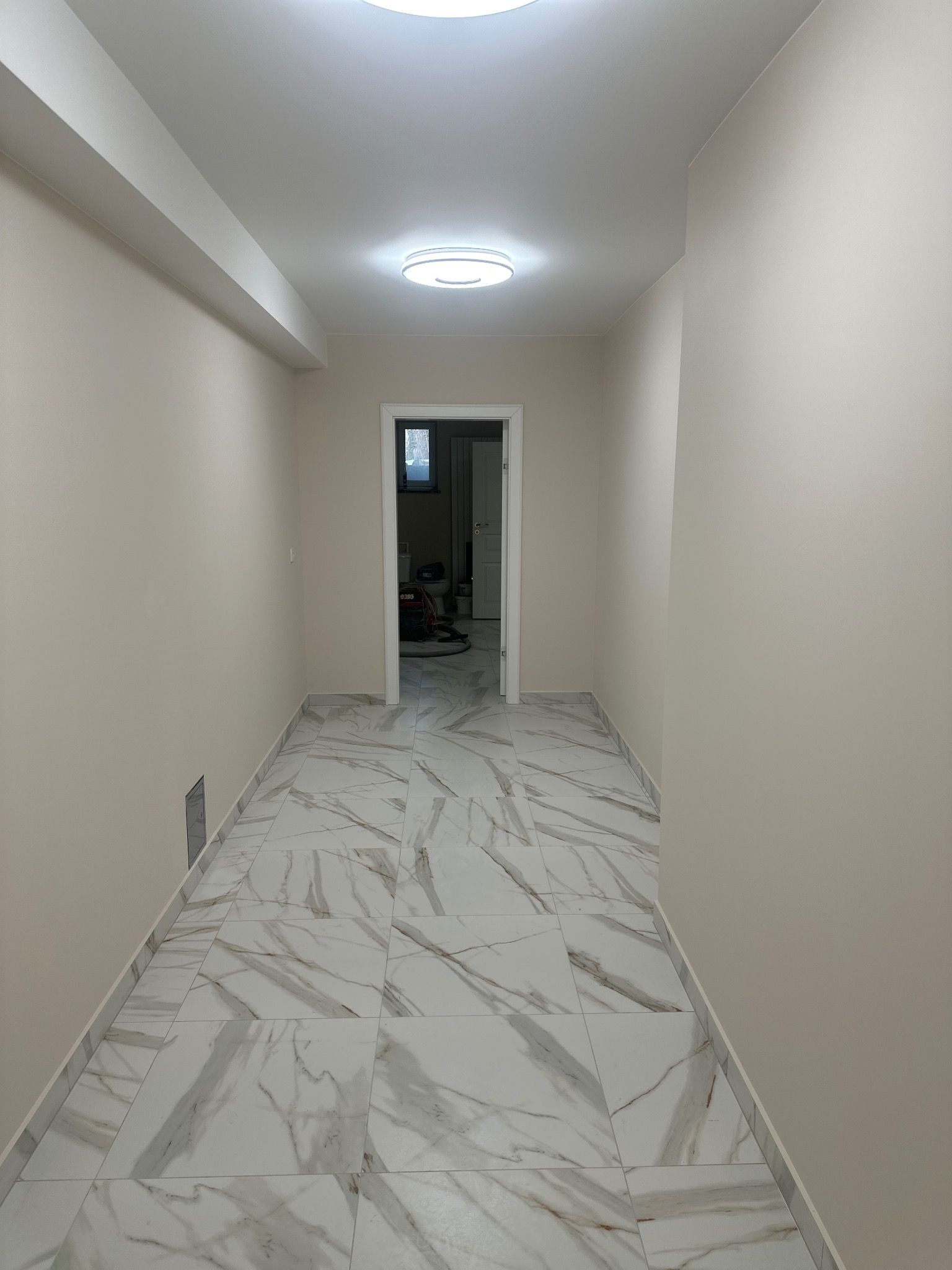 Wykończony korytarz z marmurową podłogą i świeżo pomalowanymi ścianami w odcieniach beżu. Widoczne oświetlenie sufitowe i otwarte drzwi do innego pomieszczenia.