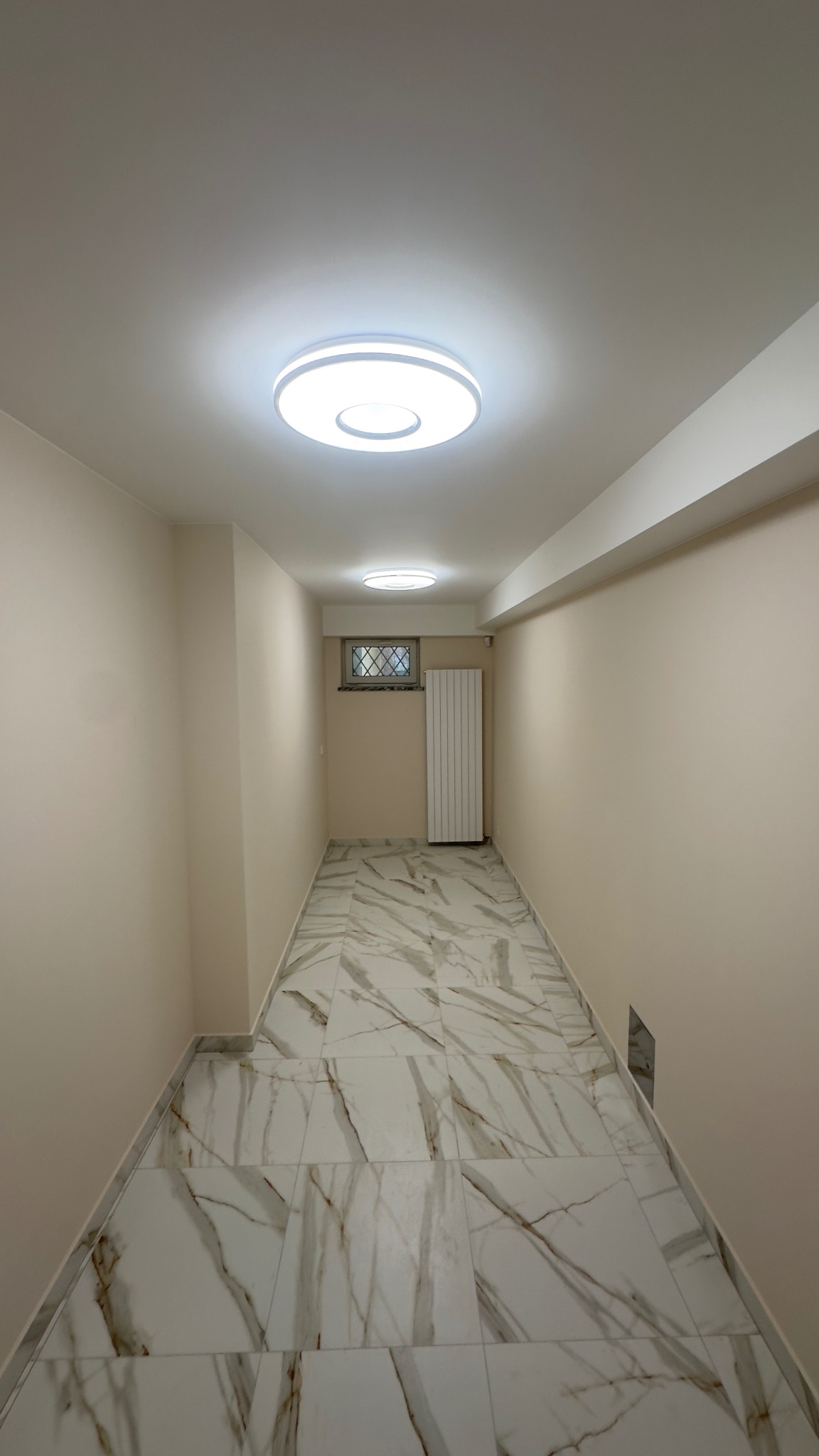 Korytarz po remoncie we Wrocławiu: marmurowe płytki podłogowe z brązowymi żyłami, kremowe ściany, nowoczesne oświetlenie sufitowe i grzejnik. Minimalistyczny design.