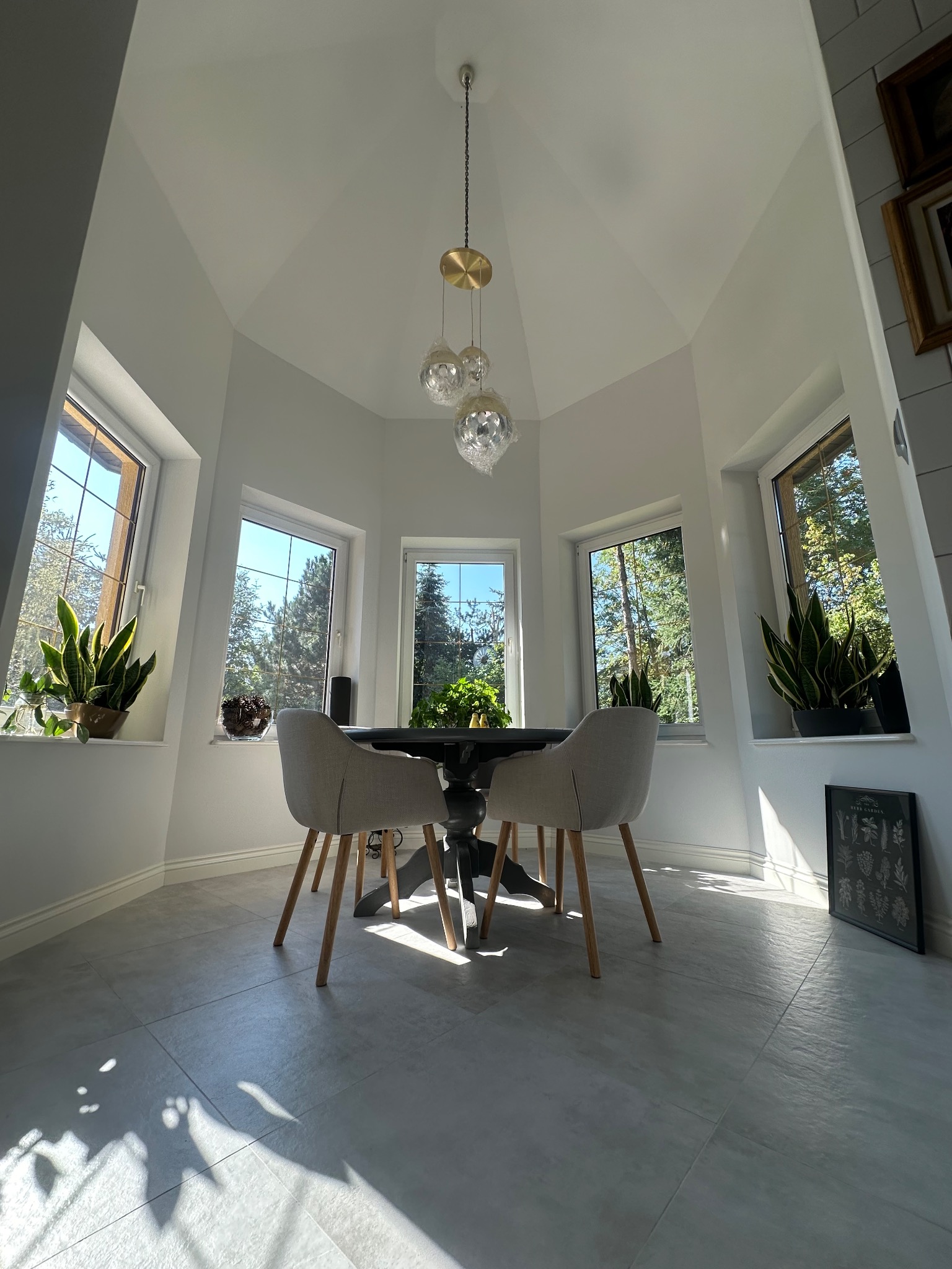 Jasne wnętrze z oknami, okrągłym stołem i krzesłami. Na parapecie rośliny doniczkowe. Na podłodze szare płytki. Nad stołem wisi lampa. Widok na zieleń za oknem.