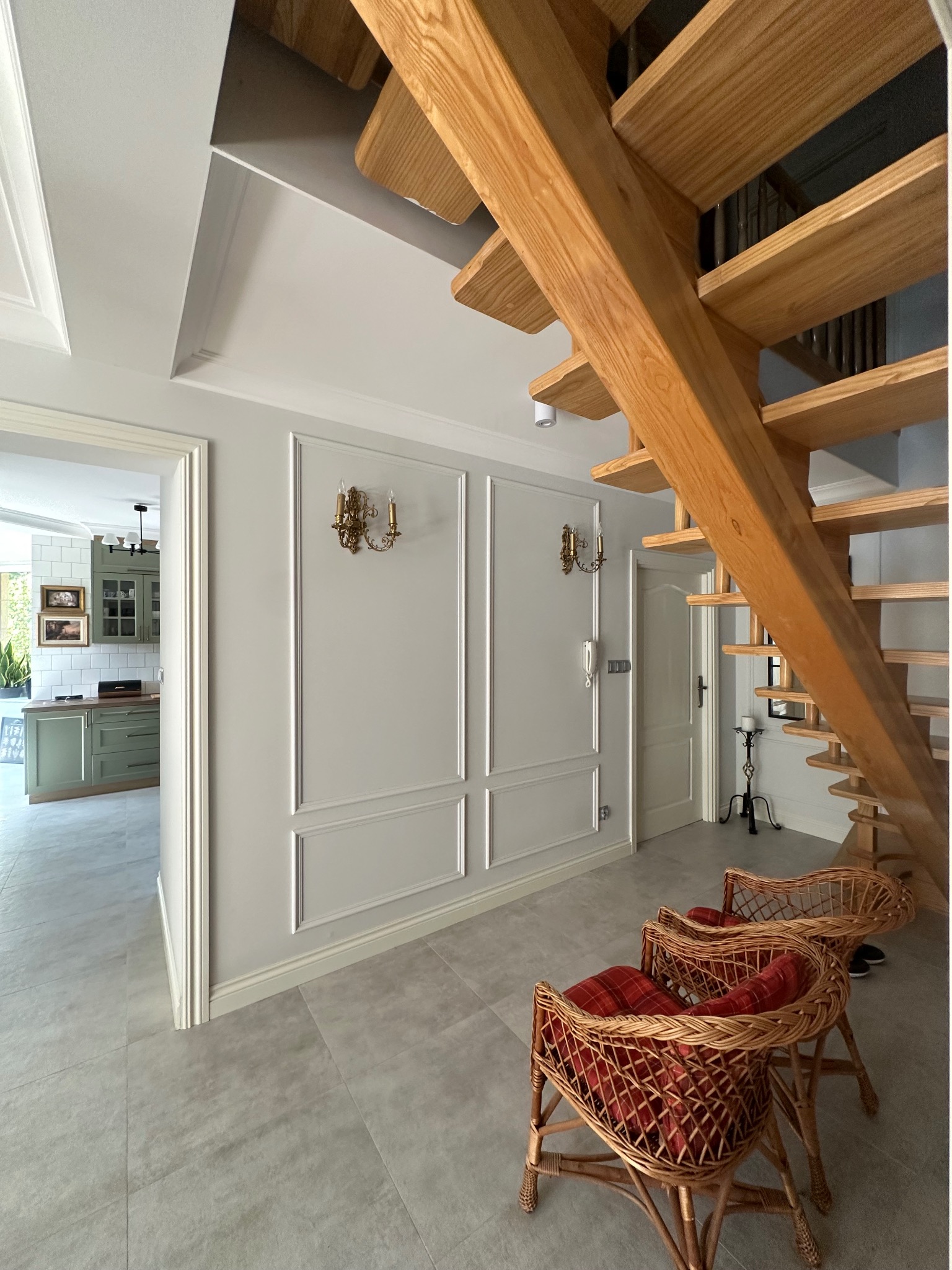 Jasne wnętrze domu z drewnianymi schodami, szarą podłogą i ścianami z dekoracyjnymi listwami. W tle widoczna kuchnia w odcieniach zieleni. Dwa wiklinowe krzesła z czerwonymi poduszkami.