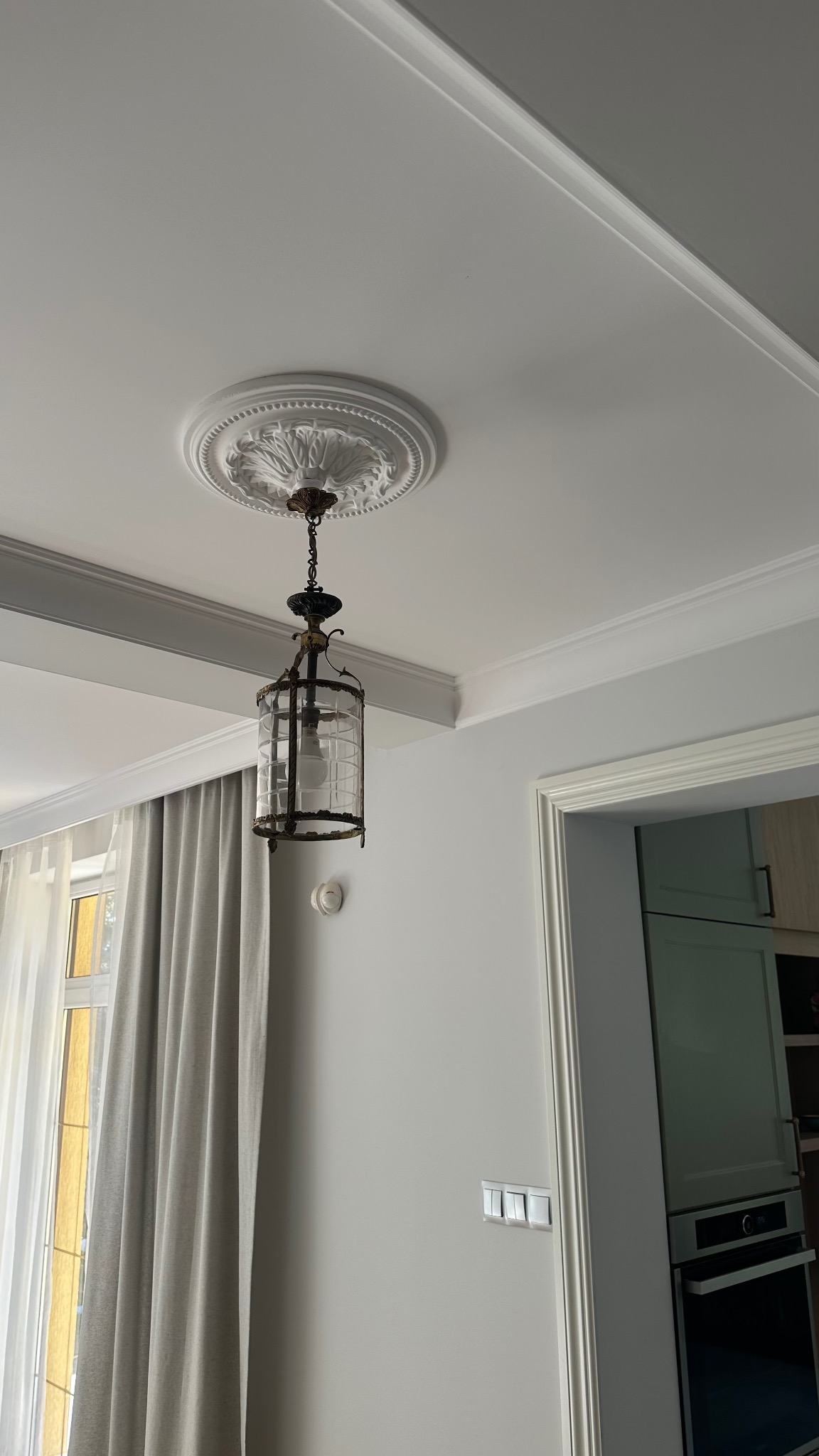 Eleganckie wykończenie sufitu z dekoracyjnym gzymsem i rozetą, na której zawieszona jest ozdobna, szklana lampa w stylu retro. Ściany w odcieniach szarości, widoczne detale wnętrza.