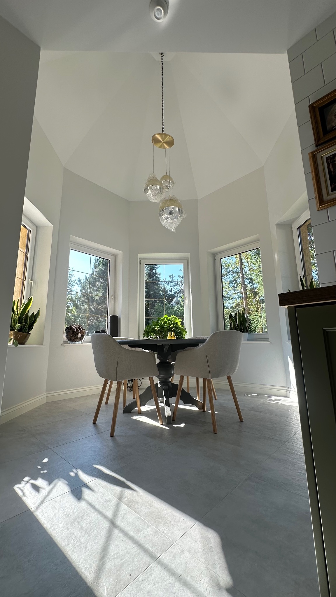 Jasne wnętrze jadalni z okrągłym stołem i krzesłami, oświetlone naturalnym światłem wpadającym przez okna. Minimalistyczny design z roślinami w doniczkach.