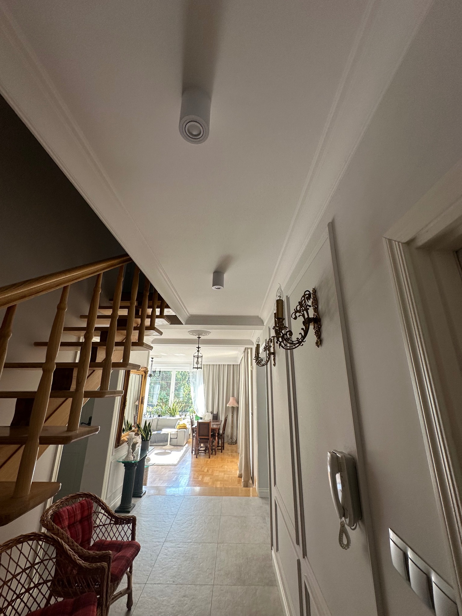 Eleganckie wykończenie wnętrza: białe ściany z dekoracyjnymi listwami, ozdobne kinkiety, schody z drewnianą balustradą i widok na salon w tle.