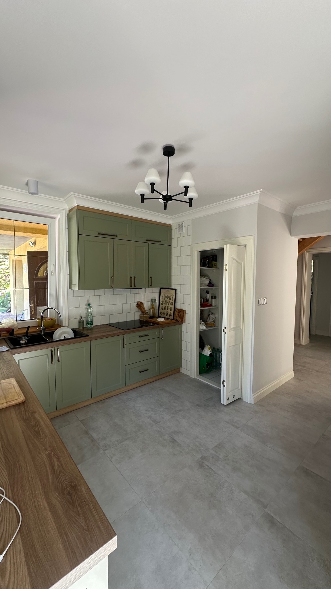 Jasna kuchnia z zielonymi szafkami i drewnianymi blatami. Widoczna spiżarnia z otwartymi drzwiami. Szara podłoga i białe ściany. Nowoczesne oświetlenie.