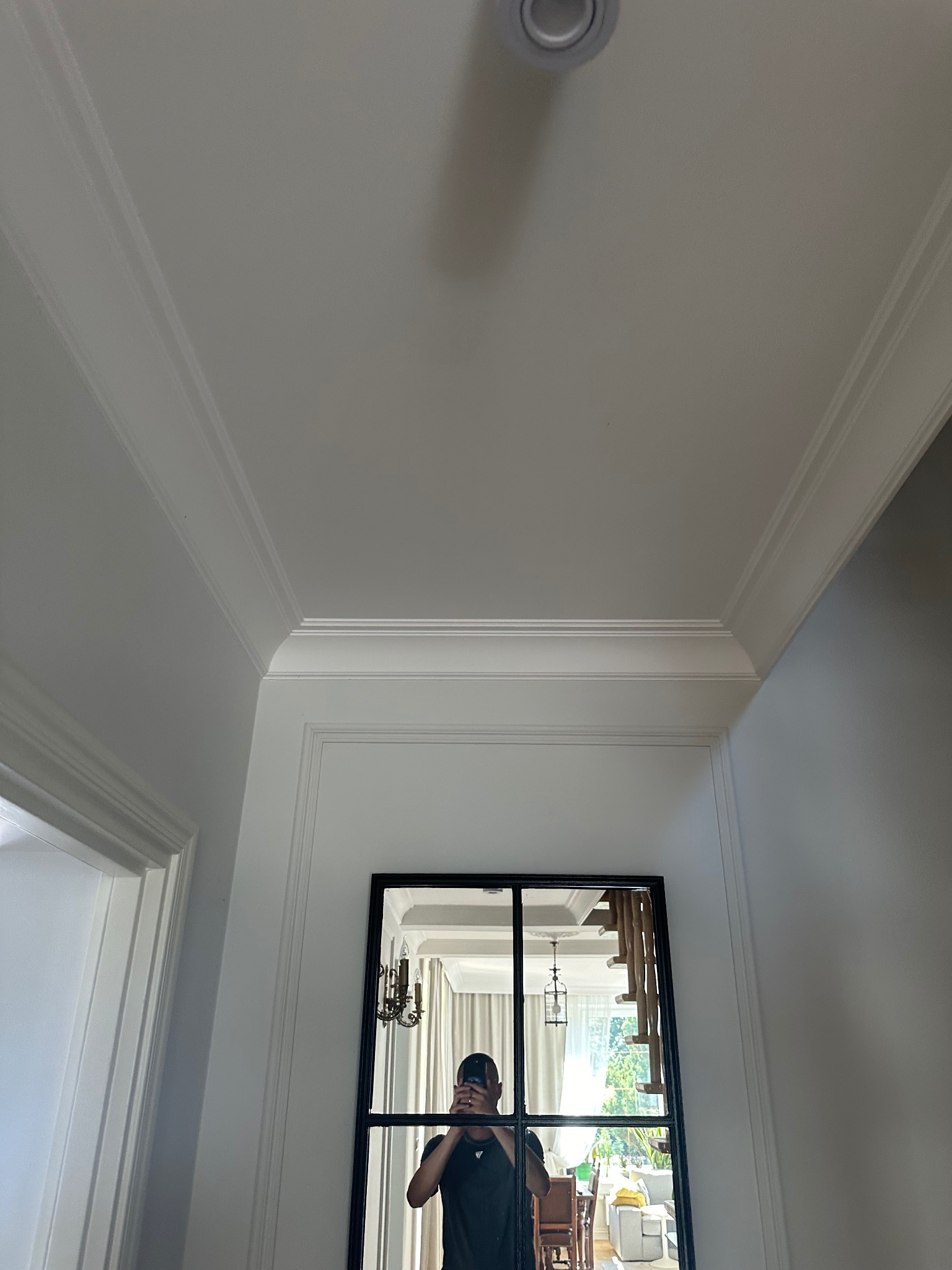 Białe, wykończone wnętrze z ozdobnymi listwami sufitowymi i ściennymi. W lustrze odbija się fragment wnętrza z schodami i osoba robiąca zdjęcie.