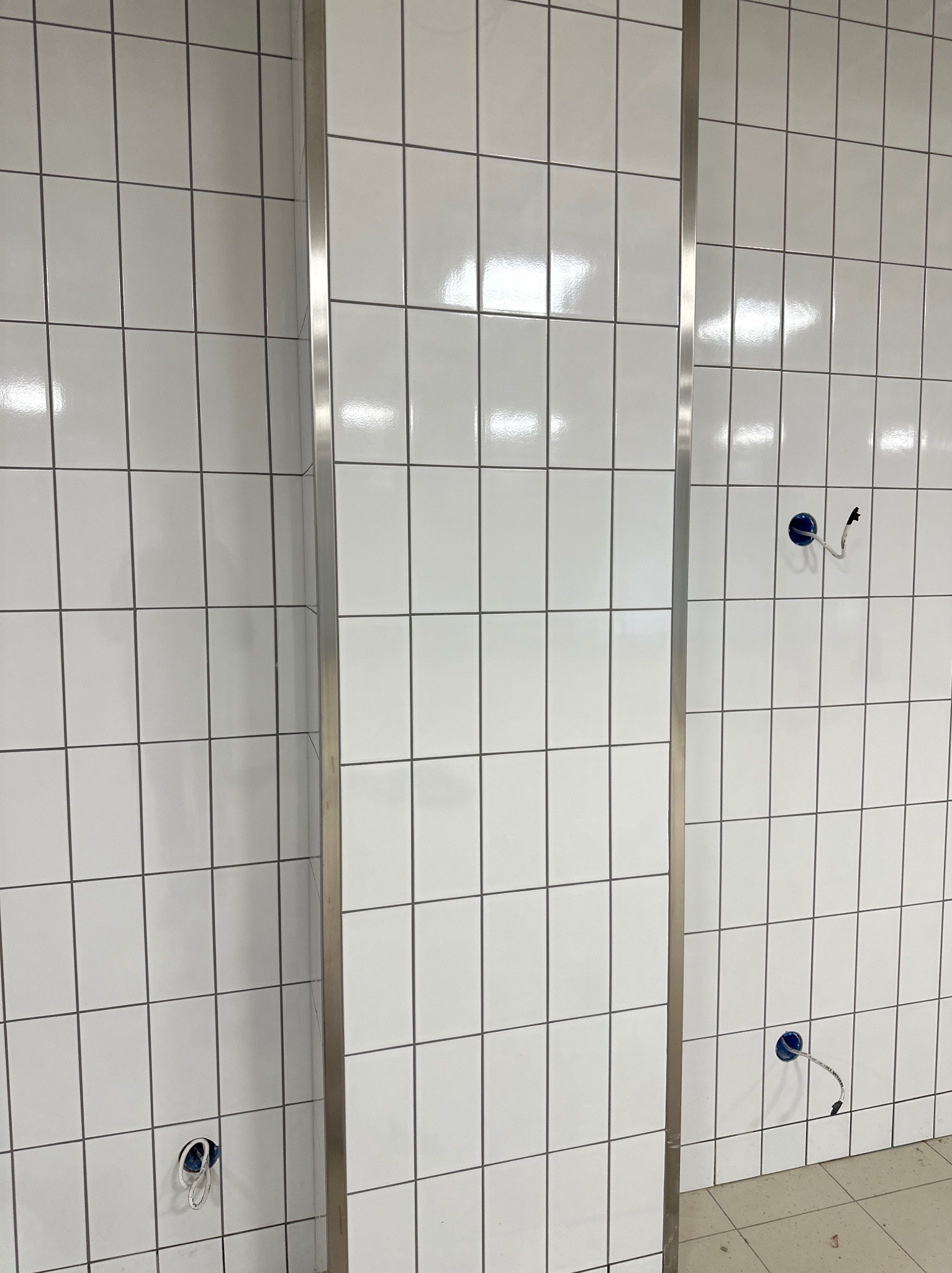 Białe, błyszczące płytki ułożone na ścianie z szarą fugą i metalową listwą. Widoczne otwory pod instalację elektryczną. Fuga epoksydowa.