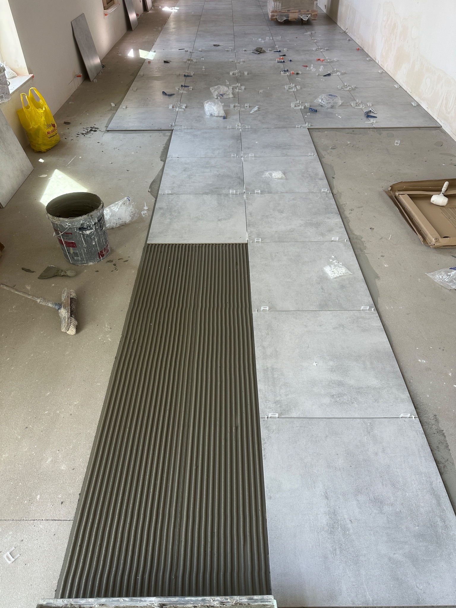 Układanie szarych, wielkoformatowych płytek na podłodze. Widoczna warstwa kleju z charakterystycznymi rowkami, krzyżyki dystansowe i narzędzia glazurnicze.