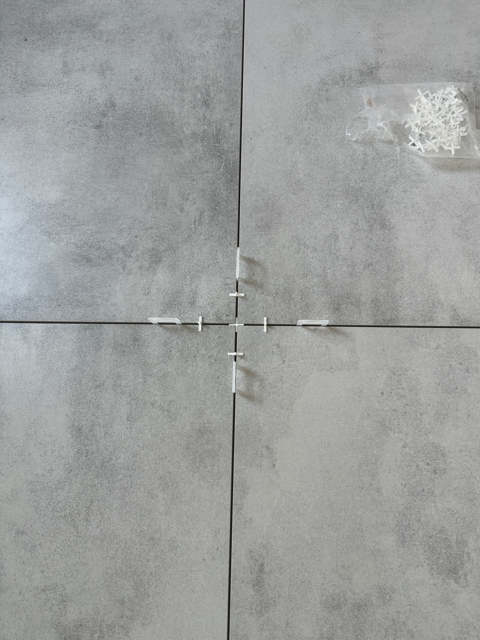 Szare płytki ułożone z krzyżykami dystansowymi, widok z góry. W rogu leży woreczek z zapasowymi krzyżykami. Minimalistyczny wzór płytek.