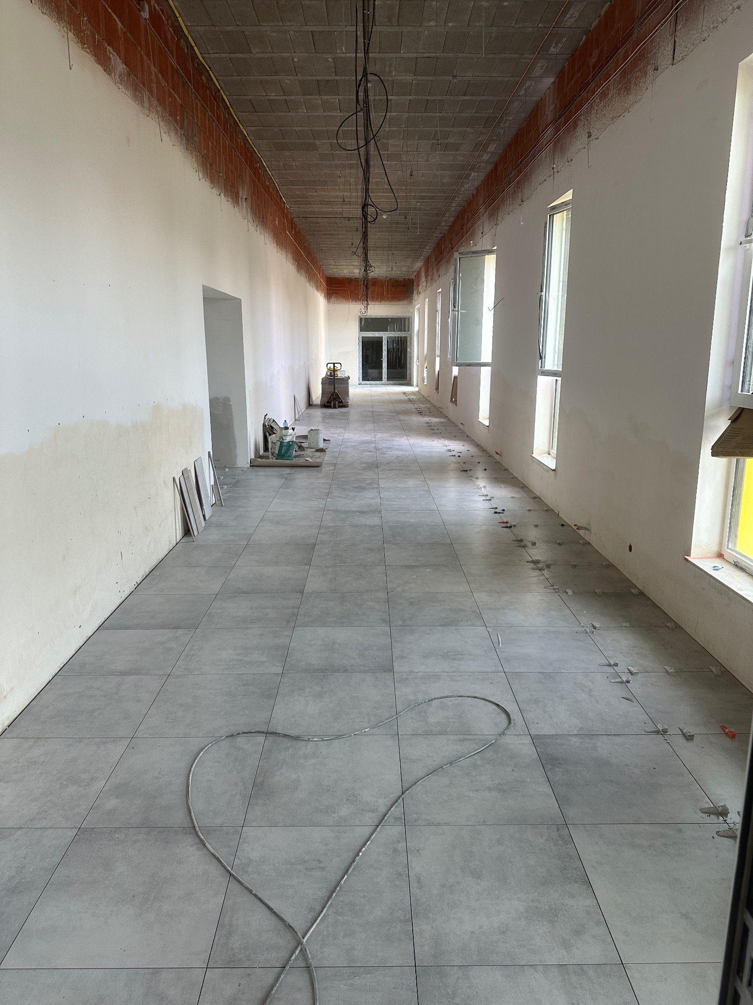 Długi korytarz w trakcie prac. Szare płytki podłogowe, ściany częściowo przygotowane do malowania, widoczne narzędzia i materiały budowlane. Surowy stan wykończenia.