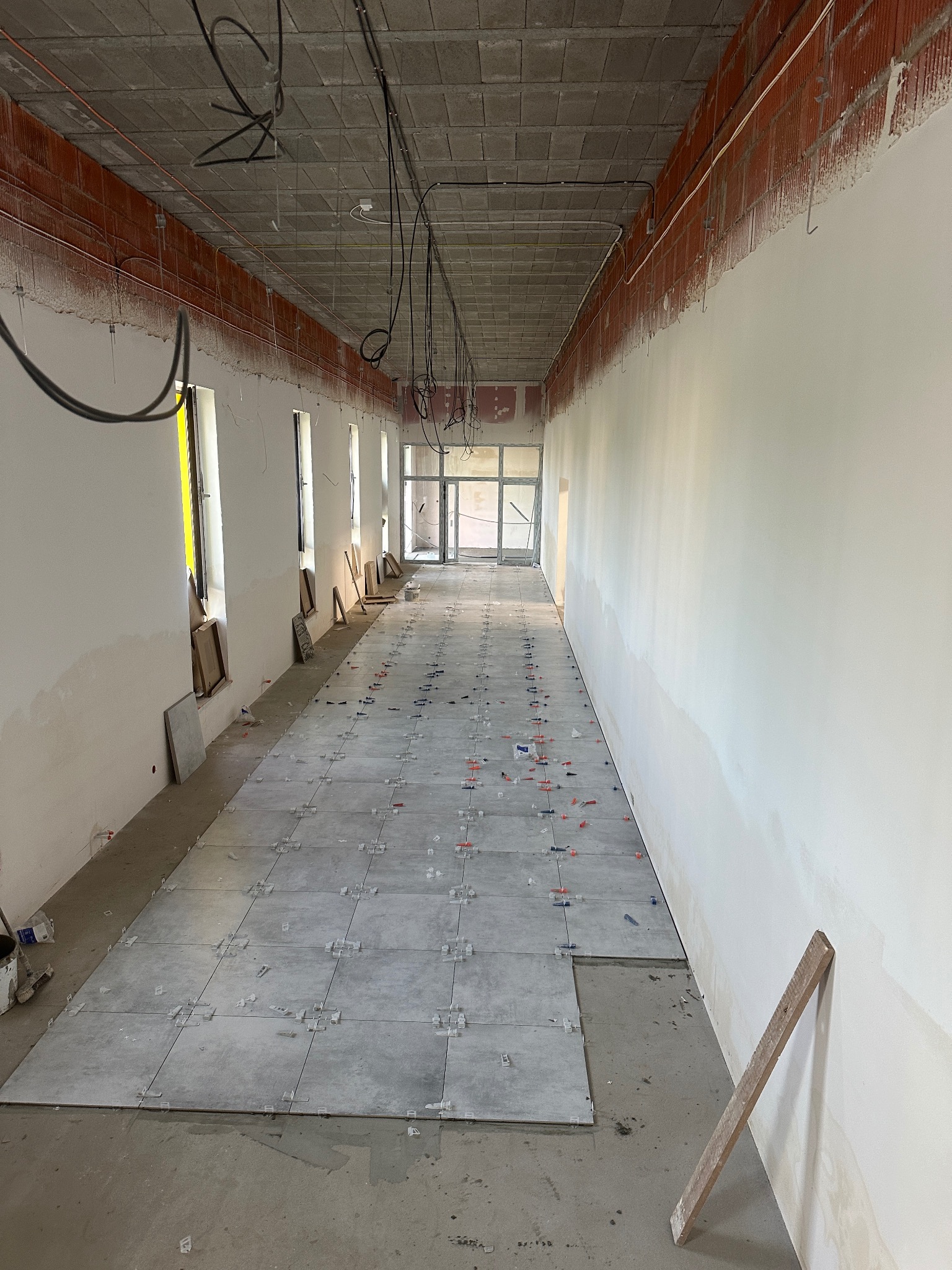 Układanie szarych płytek podłogowych w długim korytarzu, widoczne krzyżyki dystansowe, ściany przygotowane do dalszych prac, surowy sufit z instalacjami.