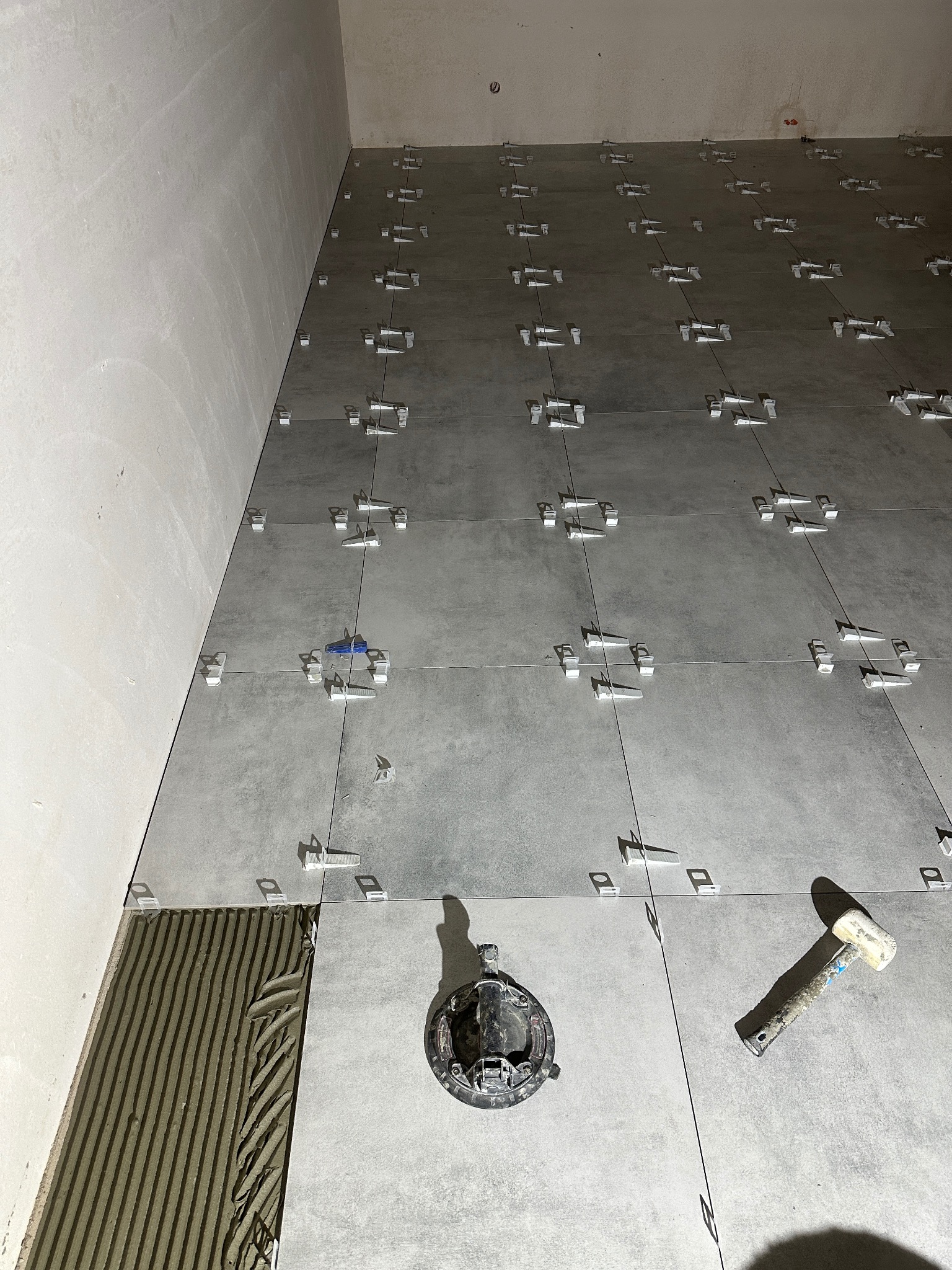 Szare płytki 60x60 ułożone na podłodze z użyciem systemu poziomowania, widoczny fragment z nałożonym klejem i gumowy młotek. Ujęcie z góry, surowy stan.