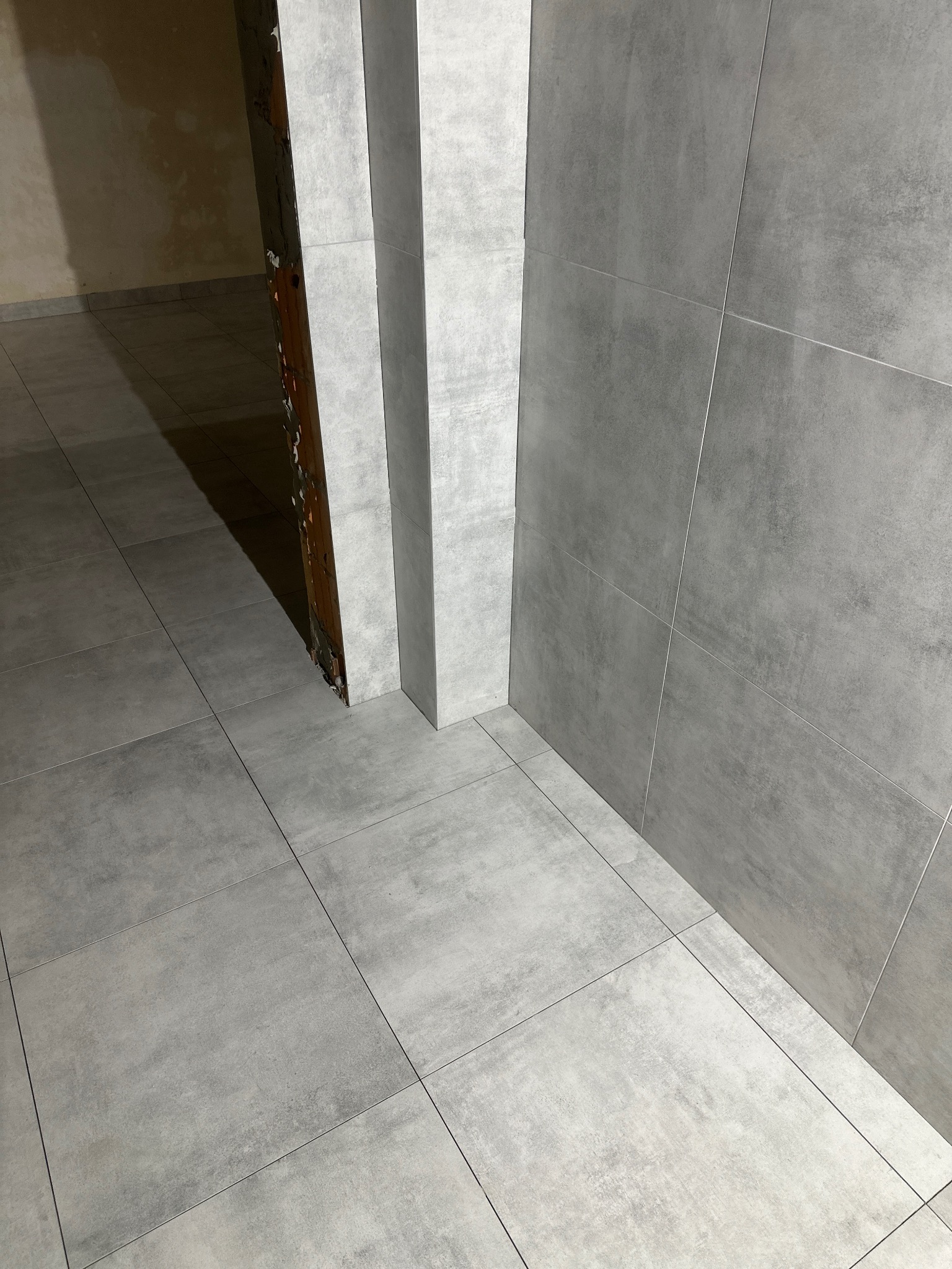 Ułożone szare płytki 60x60 na podłodze i ścianie, widoczne fugi. Fragment surowej ściany w tle. Ujęcie z niskiego kąta, podkreślające regularność wzoru.