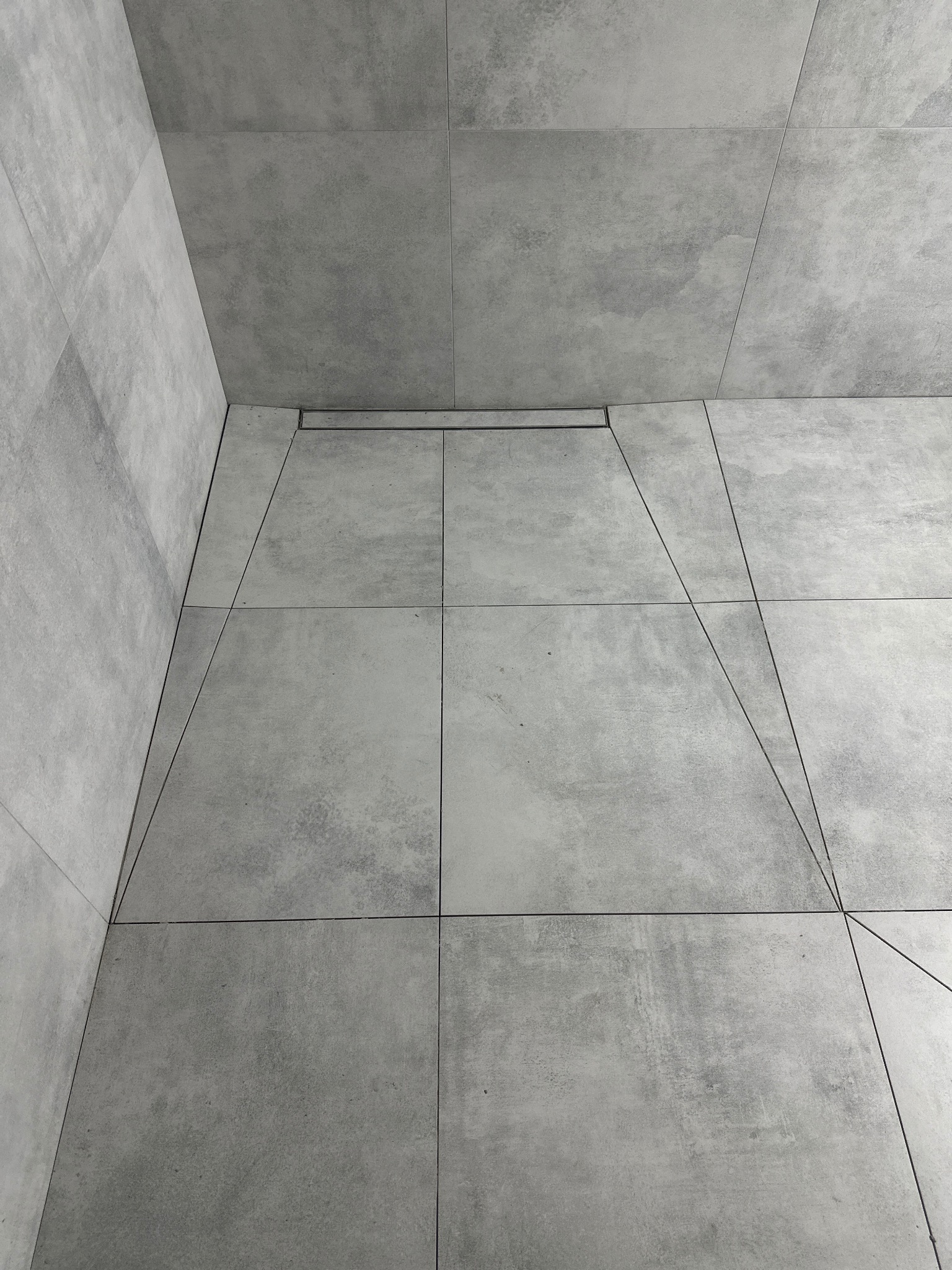 Minimalistyczna łazienka z szarymi, kwadratowymi płytkami 60x60 cm ułożonymi na podłodze i ścianach, z widocznym odpływem liniowym. Nowoczesne wykończenie.