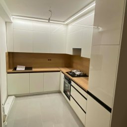 Meblosztos - Nowoczesna kuchnia z białymi, lakierowanymi frontami i drewnianym blatem. Oświetlenie LED podkreśla minimalistyczny design. Widoczna płyta grzewcza i piekarnik.