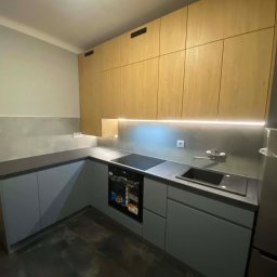 Meblosztos - Nowoczesna kuchnia na wymiar z szarymi szafkami dolnymi i drewnianymi górnymi, oświetlona listwą LED pod szafkami. Widoczny zlew, płyta indukcyjna i piekarnik.