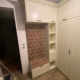 Meblosztos - Biała szafa wnękowa z tapicerowanym siedziskiem i oparciem w kolorze pudrowego różu. Widoczne półki i szuflady. Podłoga z płytek imitujących marmur.