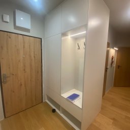 Meblosztos - Biała szafa wnękowa z otwartą przestrzenią na buty i wieszaki, oświetlona LED, w przedpokoju z drewnianą podłogą i drzwiami.