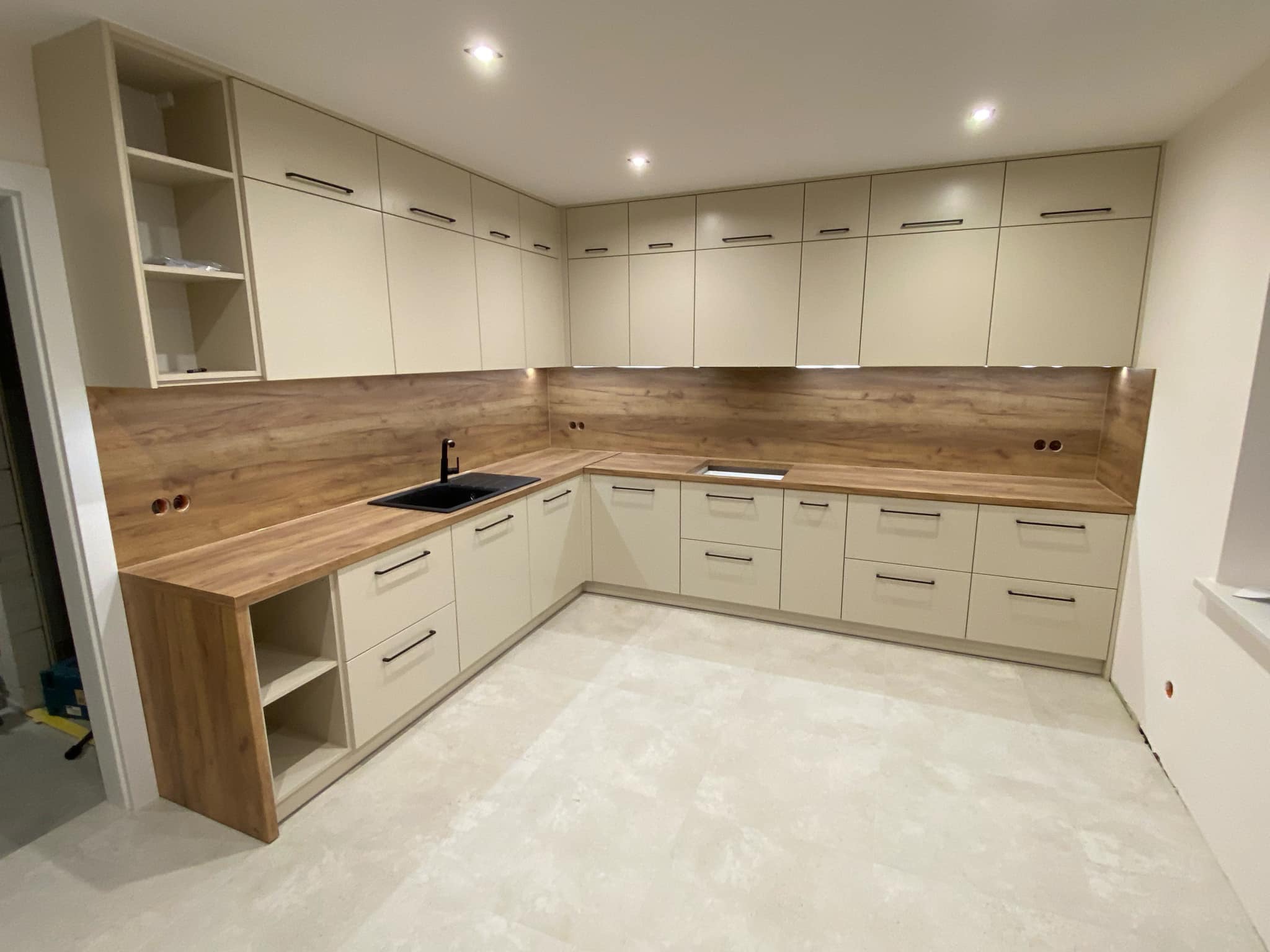 Jasna, narożna kuchnia z drewnianym blatem i czarną armaturą. Górne szafki bez uchwytów, oświetlenie punktowe. Widok na całą zabudowę, minimalistyczny styl.