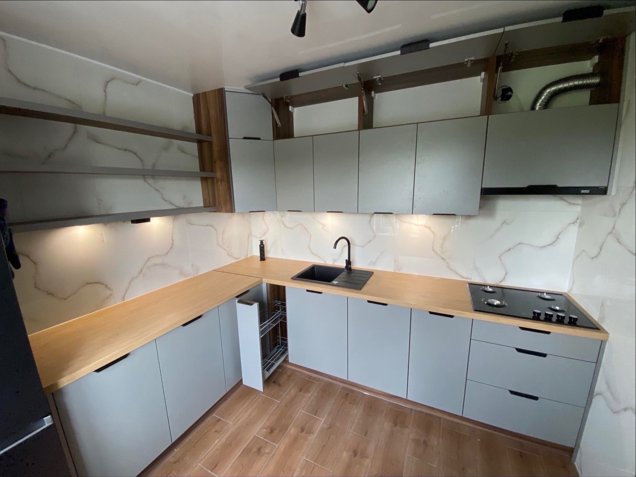 Nowoczesna kuchnia z szarymi szafkami i drewnianym blatem. Widoczne otwarte szafki górne, płyta grzewcza i wysuwany kosz cargo. Marmurowe płytki na ścianie.