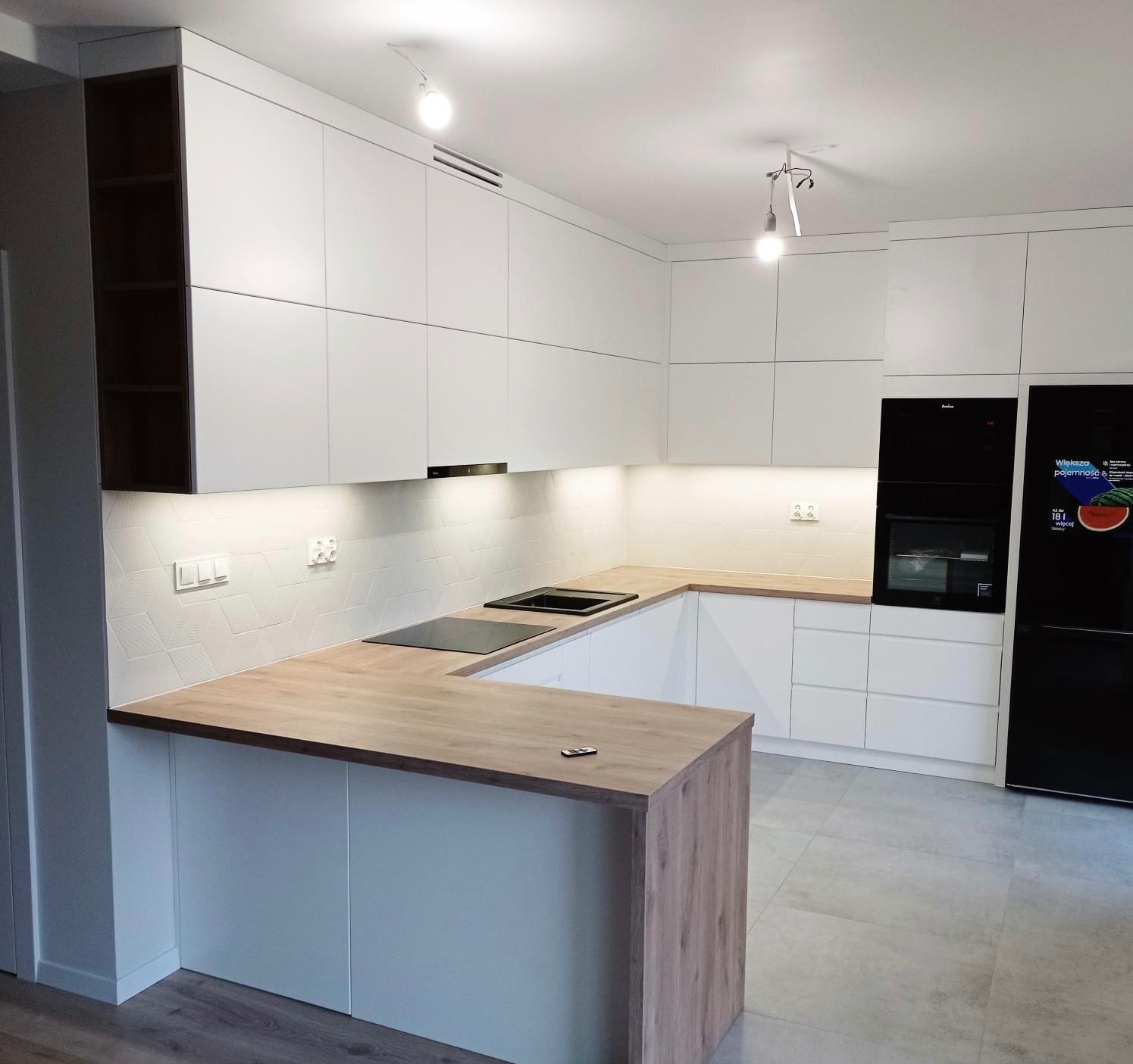 Nowoczesna, jasna kuchnia w kształcie litery U z drewnianym blatem i białymi frontami. Widoczne czarne AGD i geometryczne płytki na ścianie.