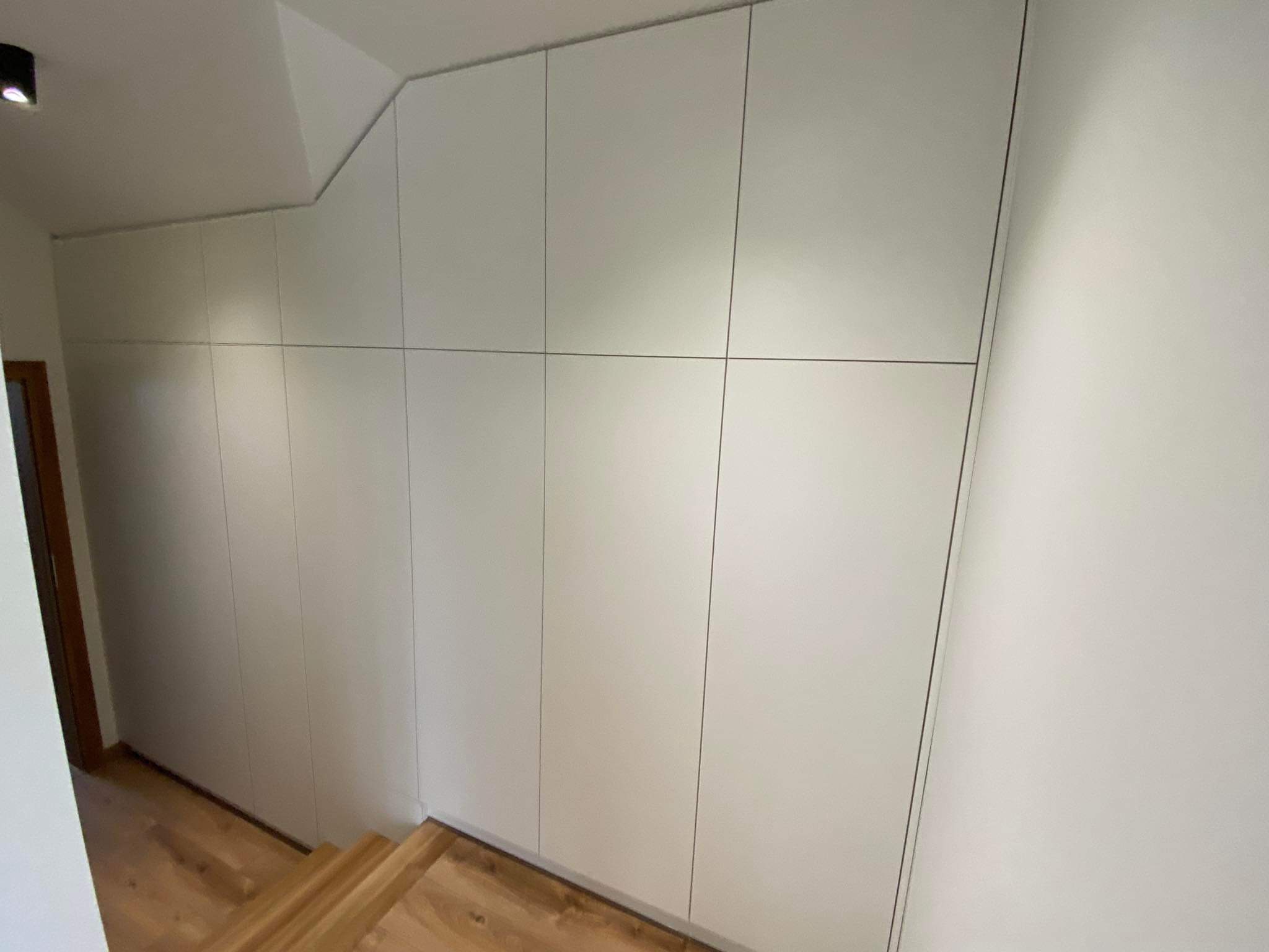 Biała szafa wnękowa na wymiar, dopasowana do skosu schodów. Minimalistyczny design, gładkie fronty bez uchwytów. Widoczne drewniane schody i fragment drzwi.