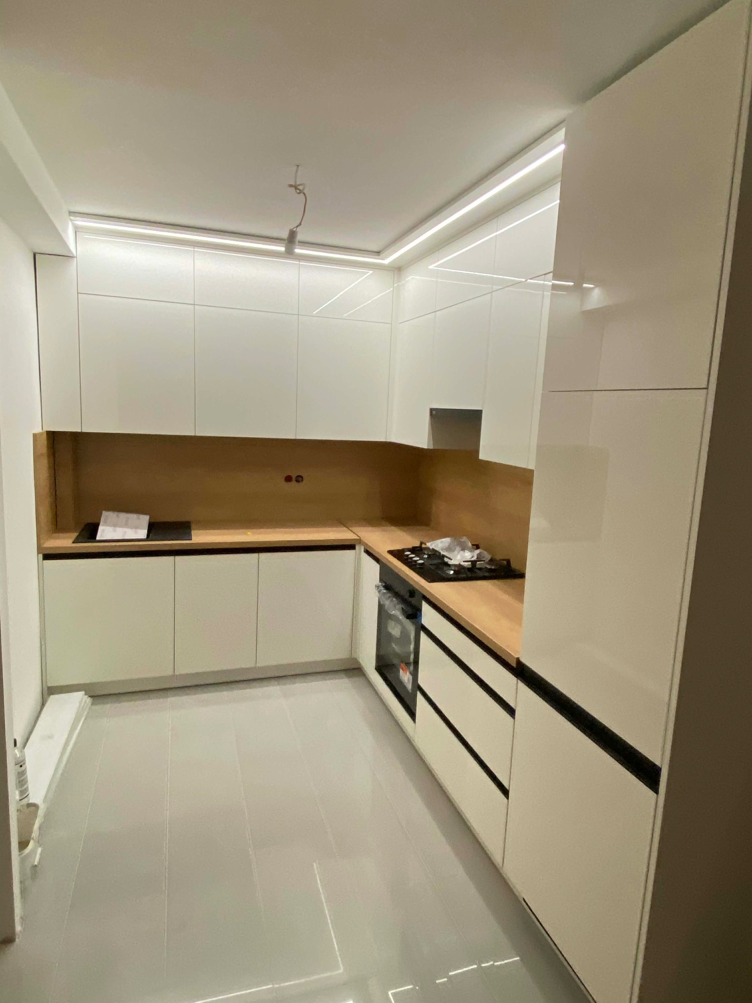 Nowoczesna kuchnia z białymi, lakierowanymi frontami i drewnianym blatem. Oświetlenie LED podkreśla minimalistyczny design. Widoczna płyta grzewcza i piekarnik.