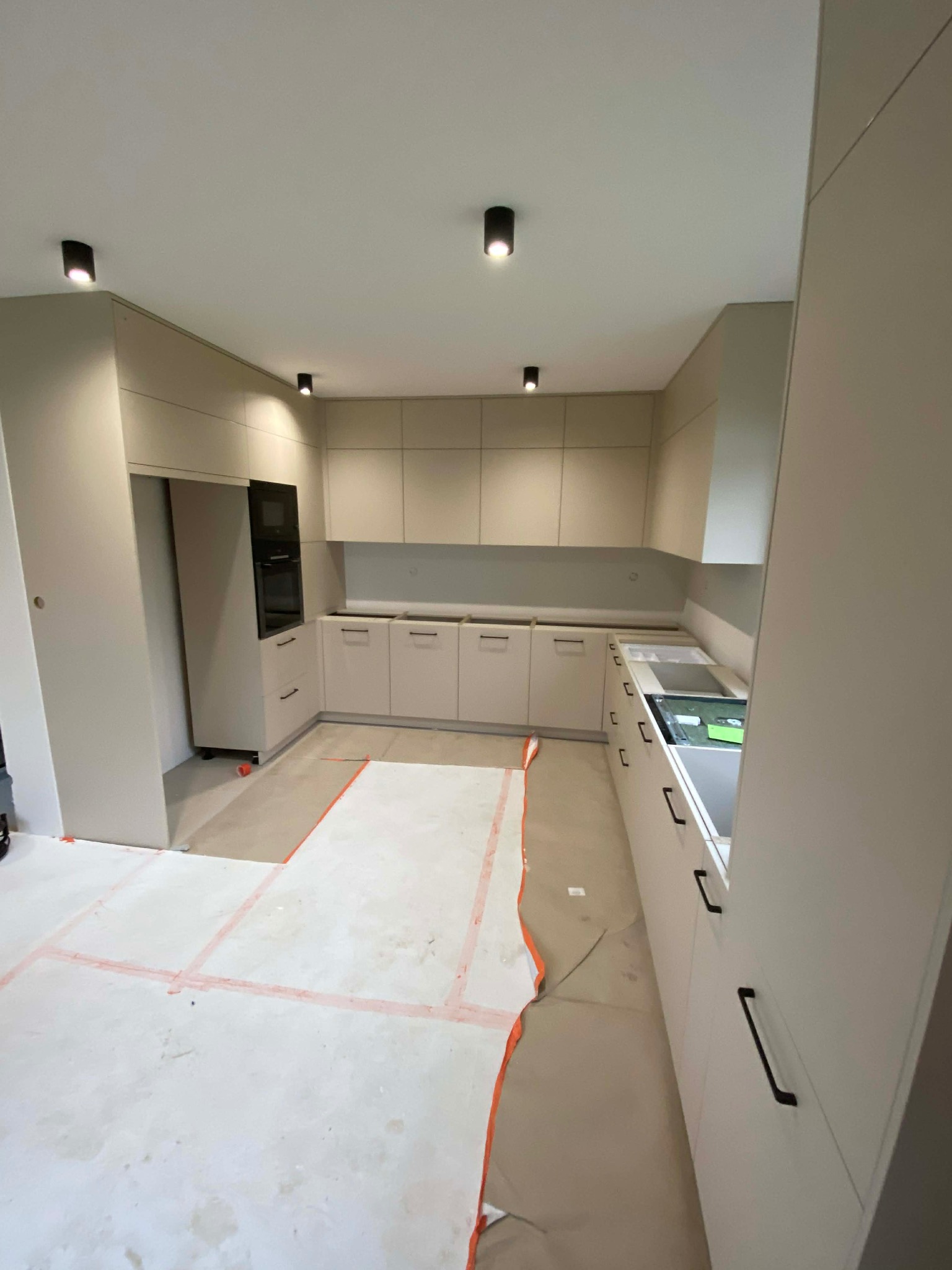 Jasna kuchnia w trakcie montażu: widoczne szafki dolne i górne, zabudowa lodówki z piekarnikiem. Podłoga zabezpieczona kartonem. Minimalistyczne oświetlenie sufitowe.