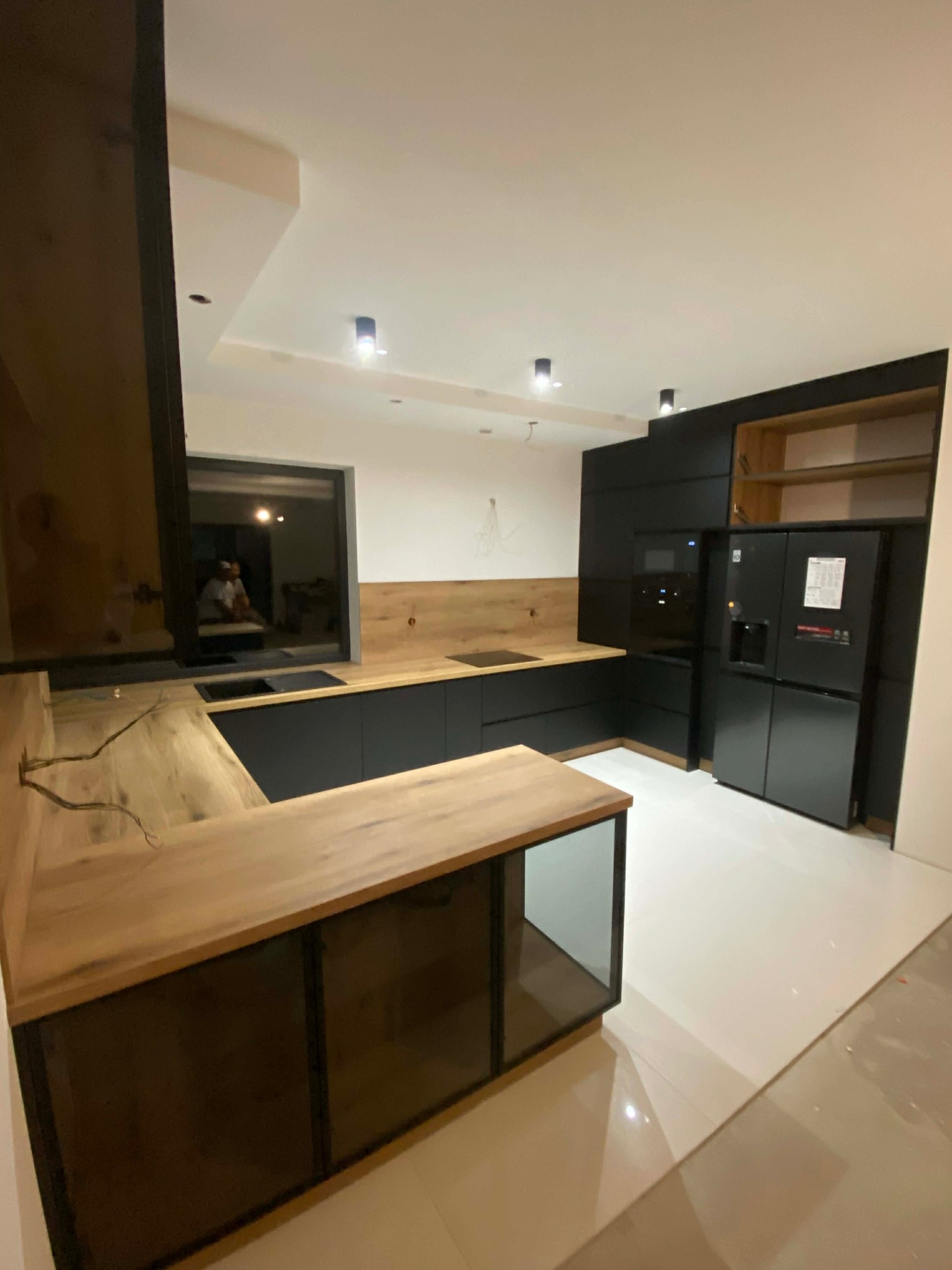 Nowoczesna kuchnia z czarnymi frontami i drewnianym blatem. Widoczna wyspa z metalową ramą i szklanymi panelami. Minimalistyczne oświetlenie punktowe.