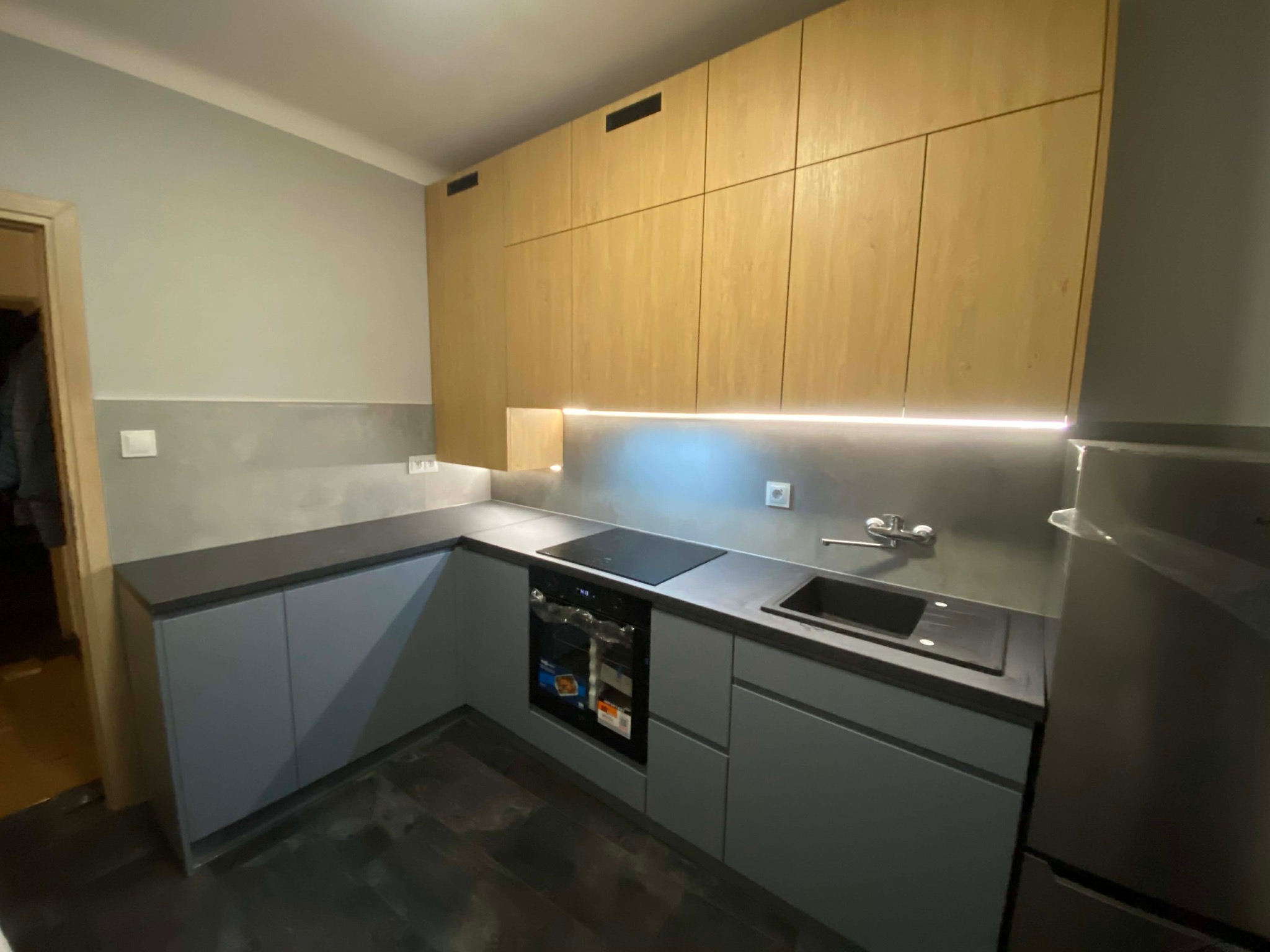 Nowoczesna kuchnia na wymiar z szarymi szafkami dolnymi i drewnianymi górnymi, oświetlona listwą LED pod szafkami. Widoczny zlew, płyta indukcyjna i piekarnik.